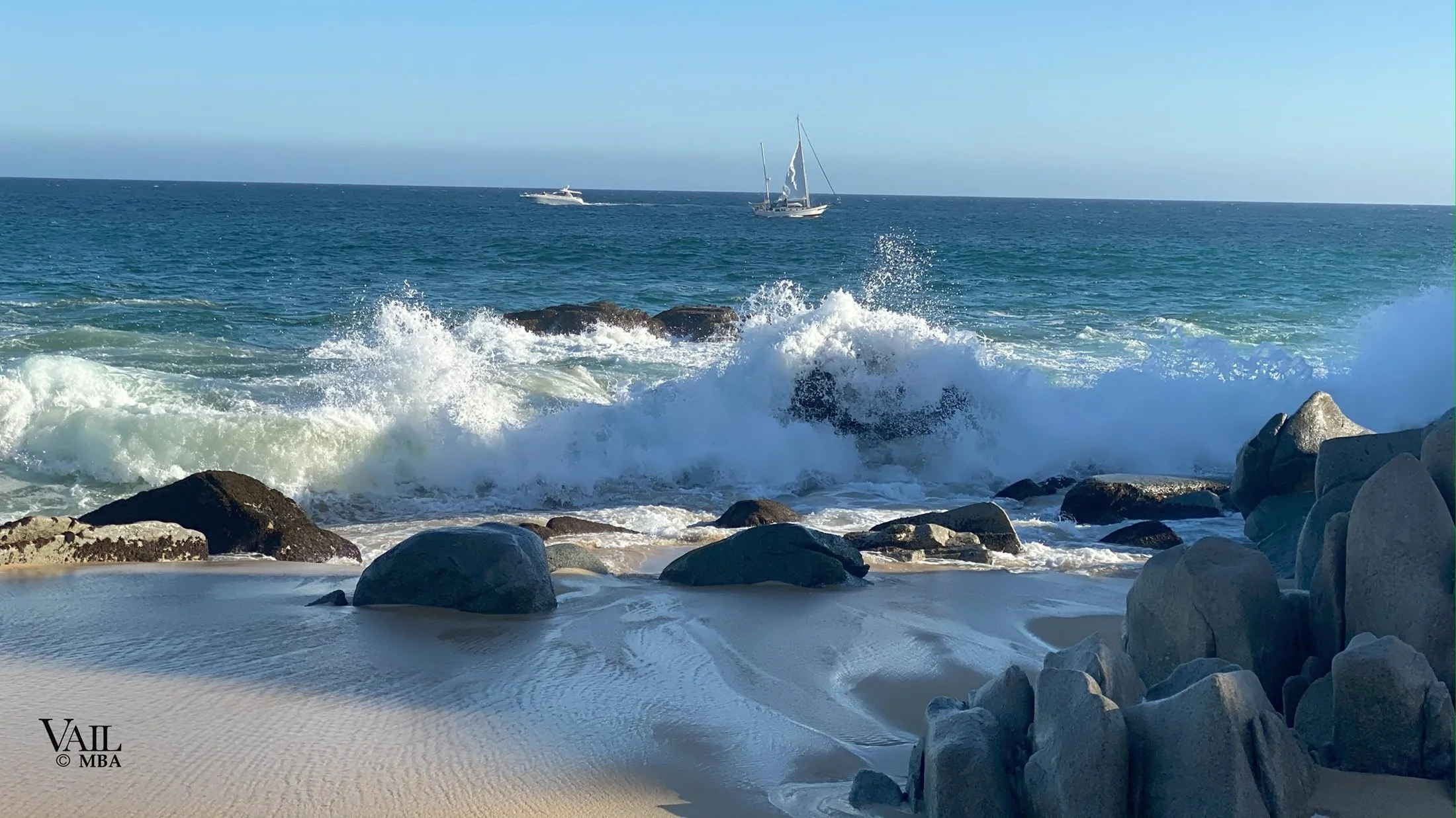 Oceanside with waves crashing on rocks and two boats sailing on the water, clear sky overhead, Cabo San Lucas.