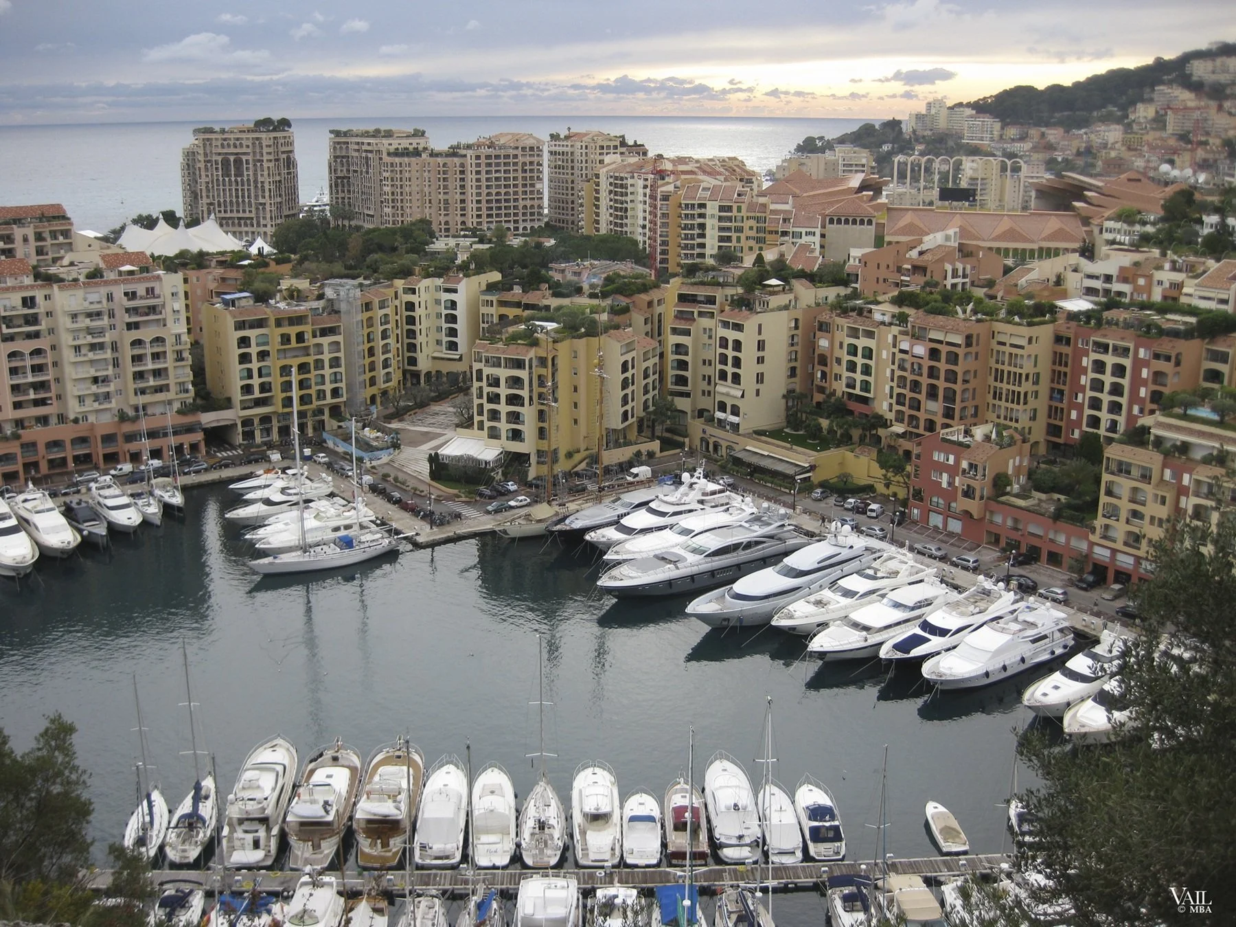 Monaco marina with boats and yachts, surrounded by high-rise buildings, and hills in the background at sunset.