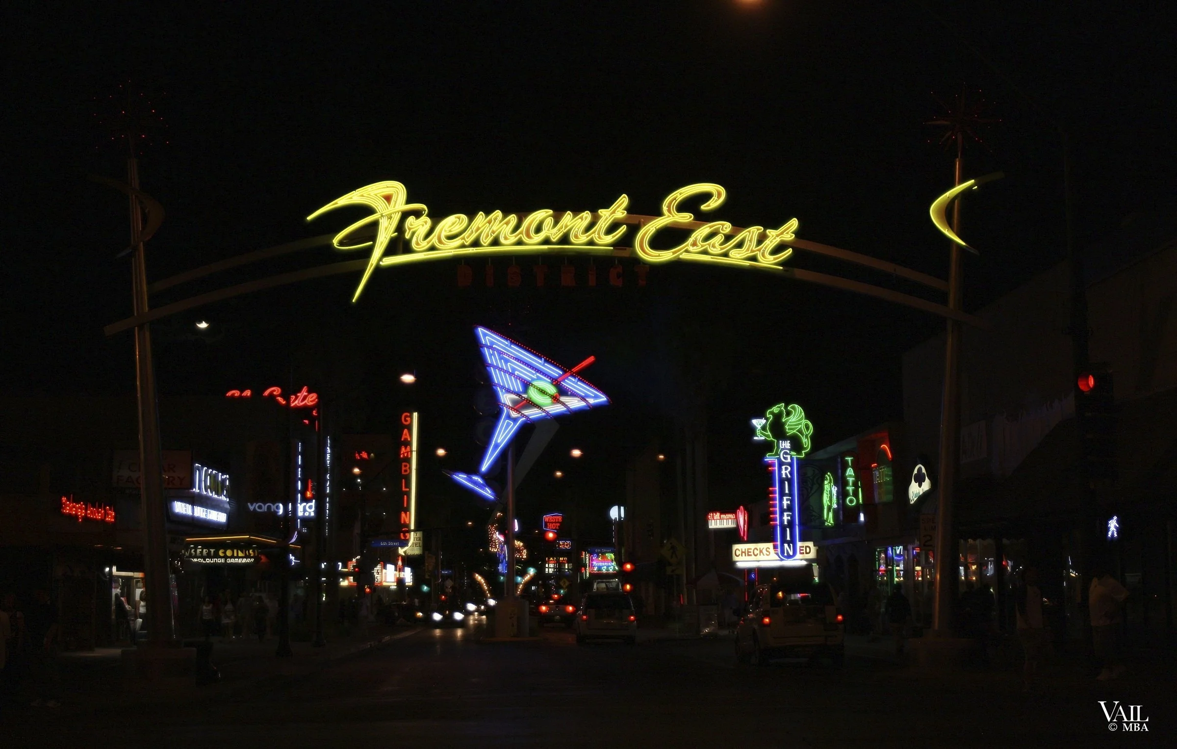 Nighttime view of Fremont East entertainment district with neon signs, including a large yellow neon sign that reads 'Fremont East' and various smaller colorful neon signs of bars, clubs, and other venues.