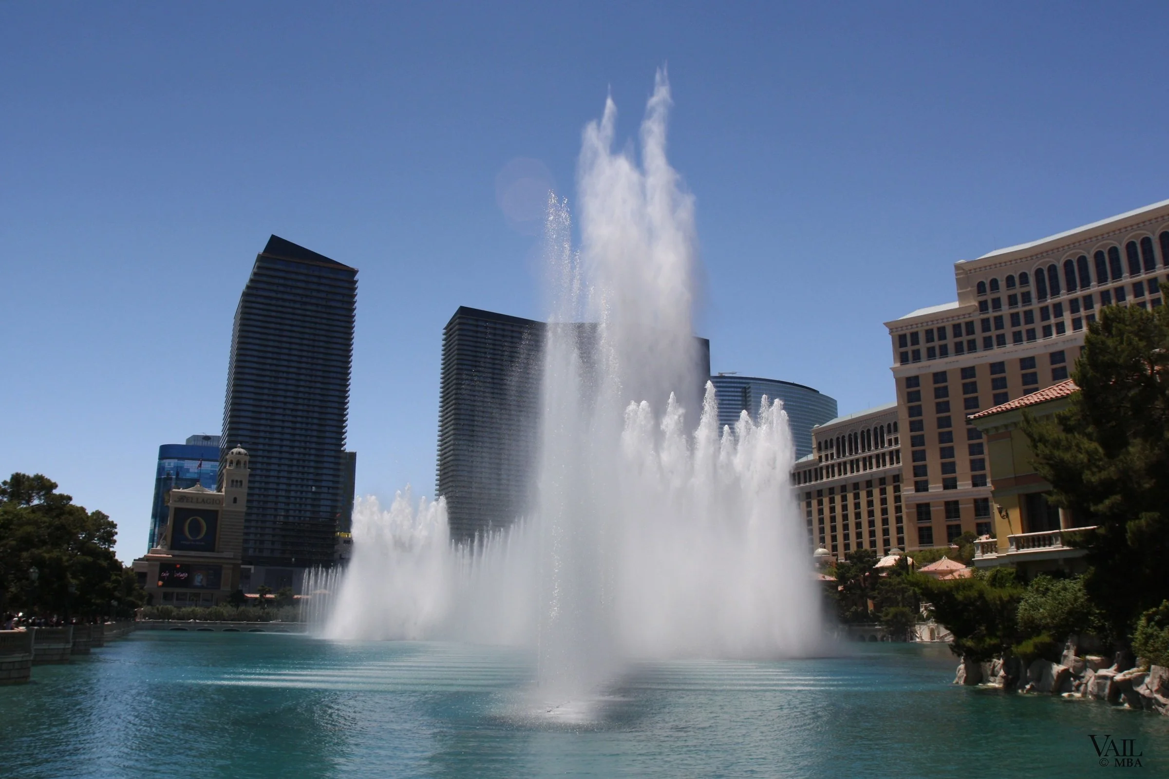 Day photo of the famous Fountains of Bellagio in Las Vegas, with water jets shooting high into the sky against a backdrop of modern skyscrapers and buildings, under a clear blue sky.