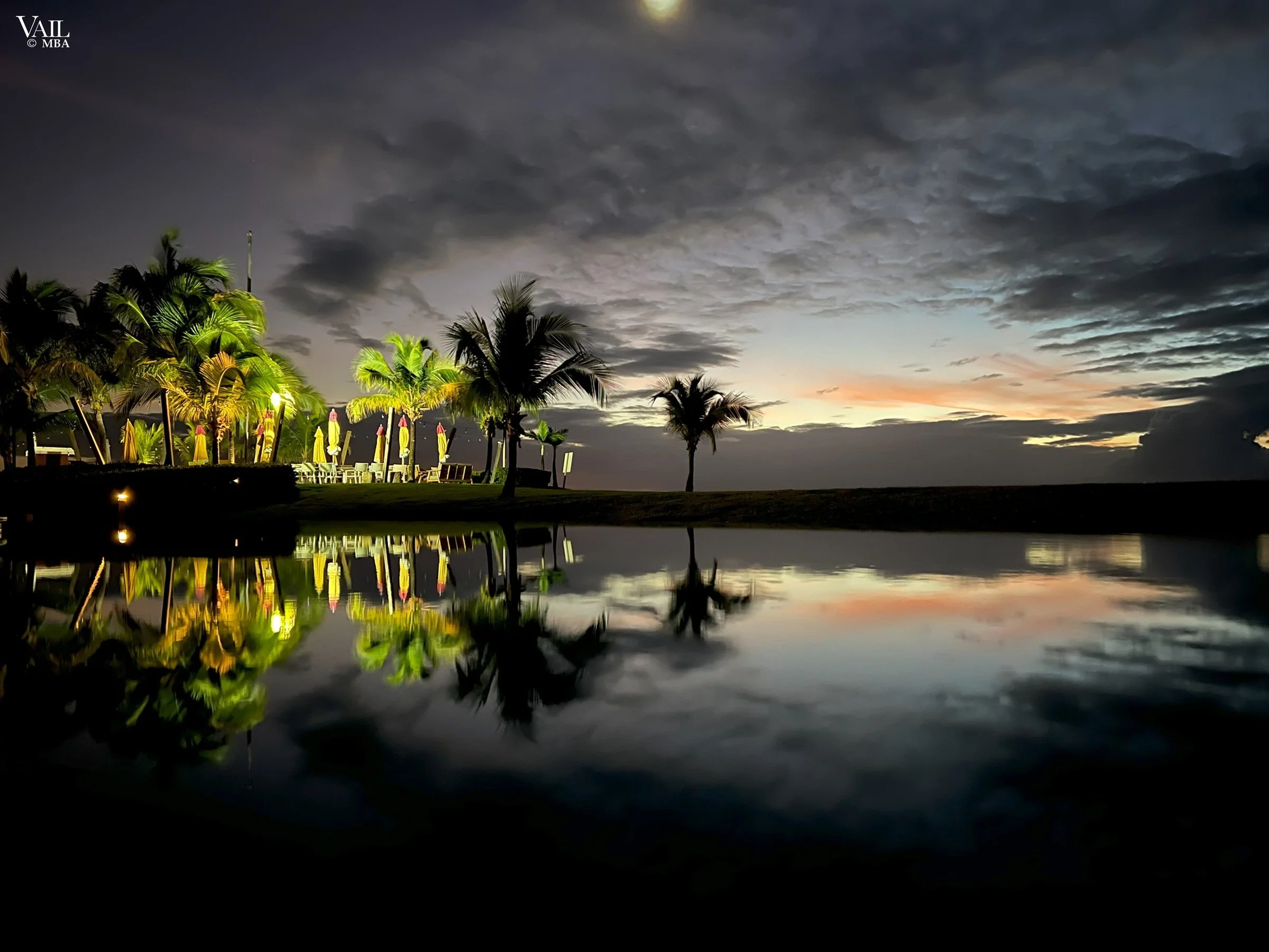 A tropical resort scene at sunset with palm trees, lounge chairs with umbrellas, and a reflective pool mirroring the darkening sky with clouds, Four Seasons Nevis.