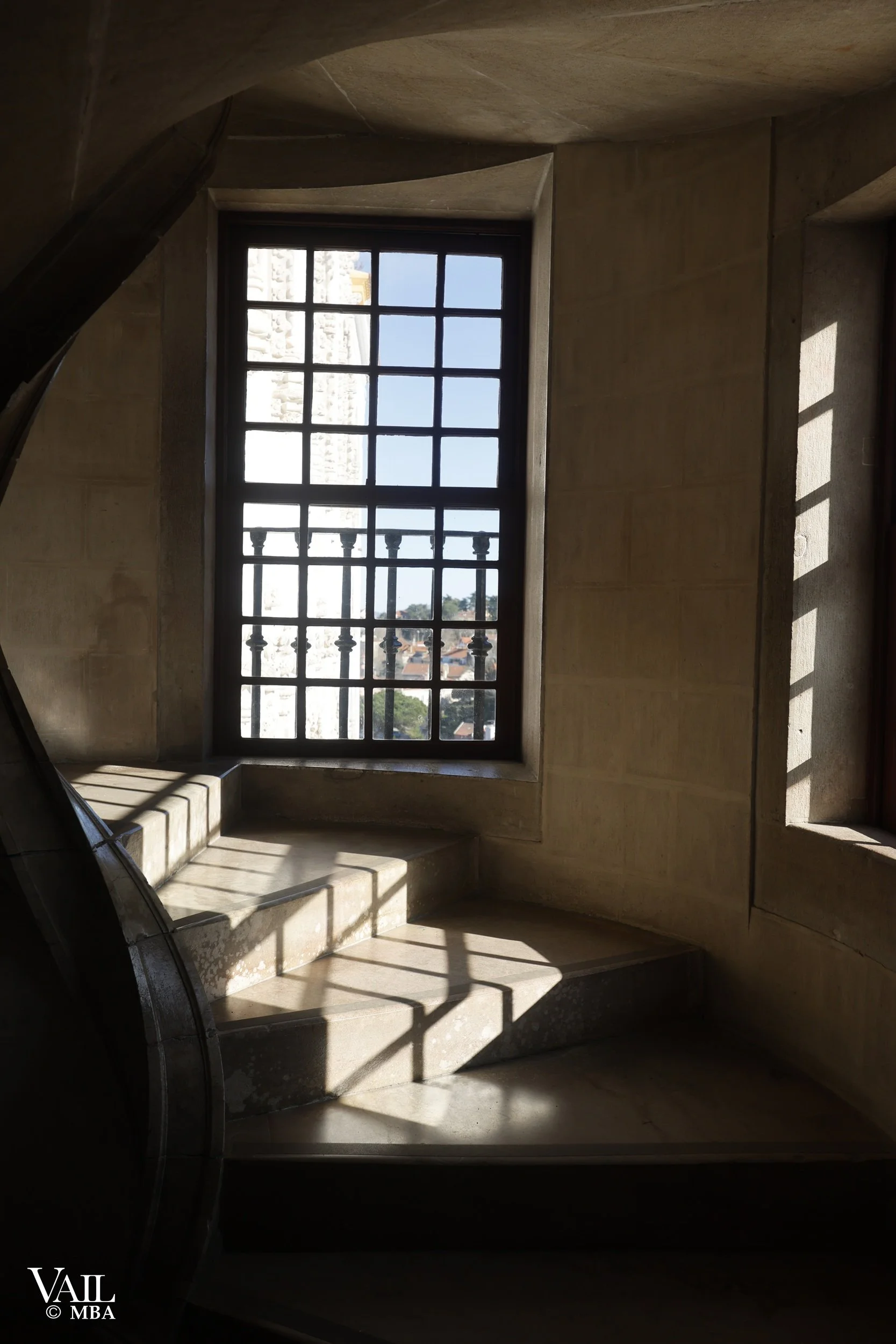 Castle staircase with sunlight casting shadows on the steps, facing a large window with a view of a cityscape and a tower outside, in Portugal.