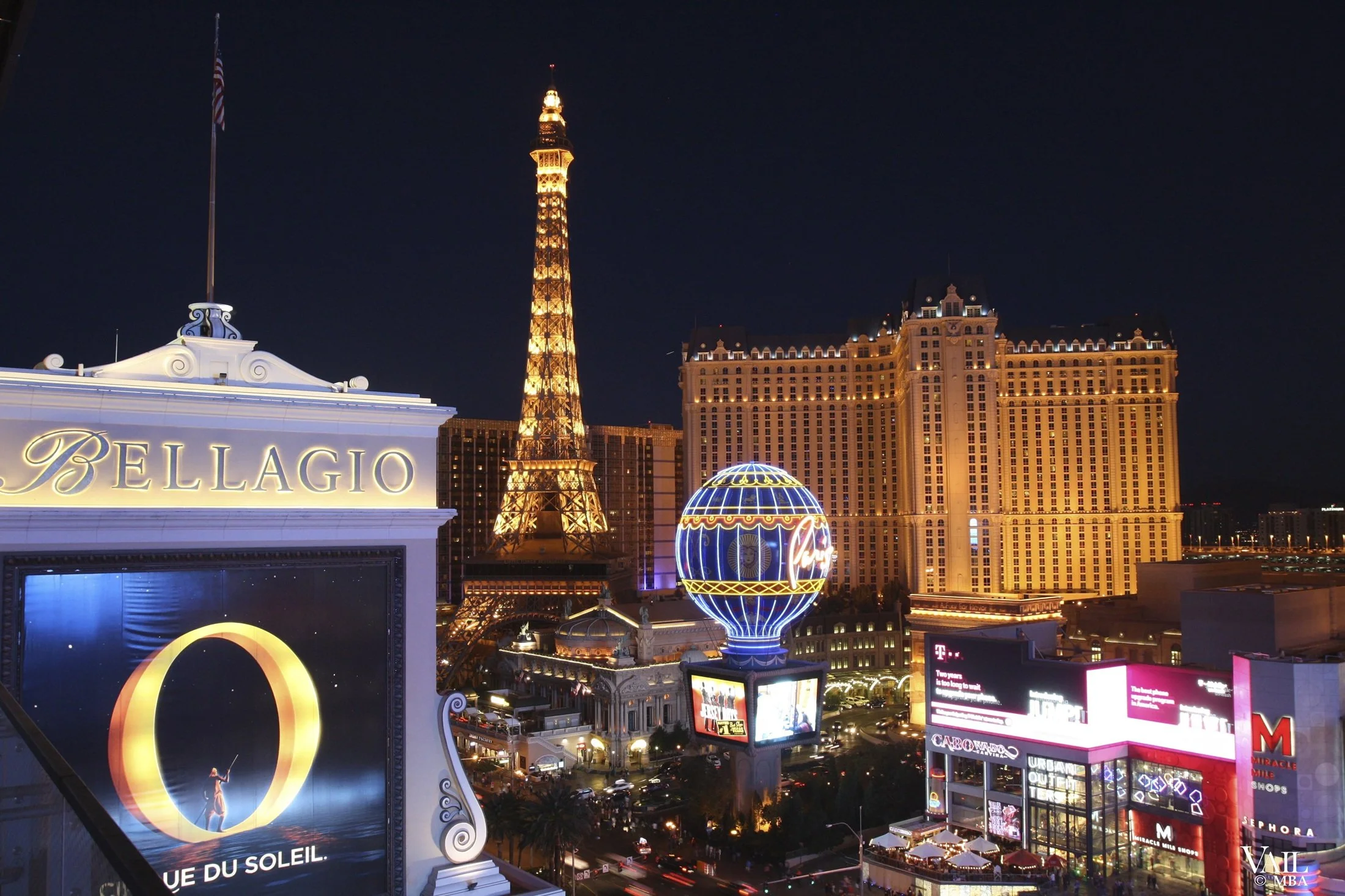 Night view of Las Vegas Strip with illuminated hotel buildings, colorful billboards, and signs including Bellagio, and Paris Las Vegas with its iconic globe and Eiffel Tower replica.