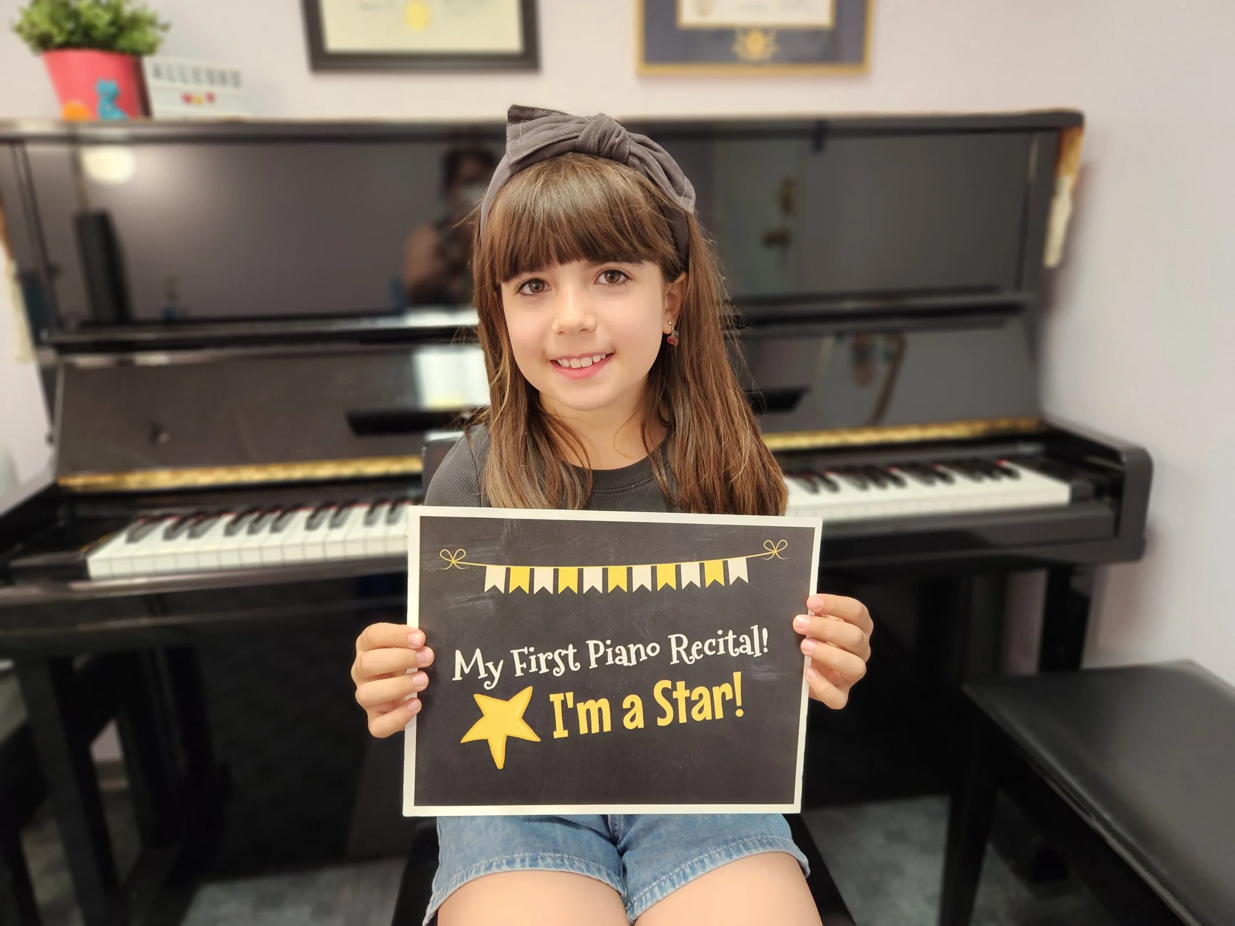Smiling girl sits in front of piano holding sign that says "My First Piano Recital I'm a Star!"
