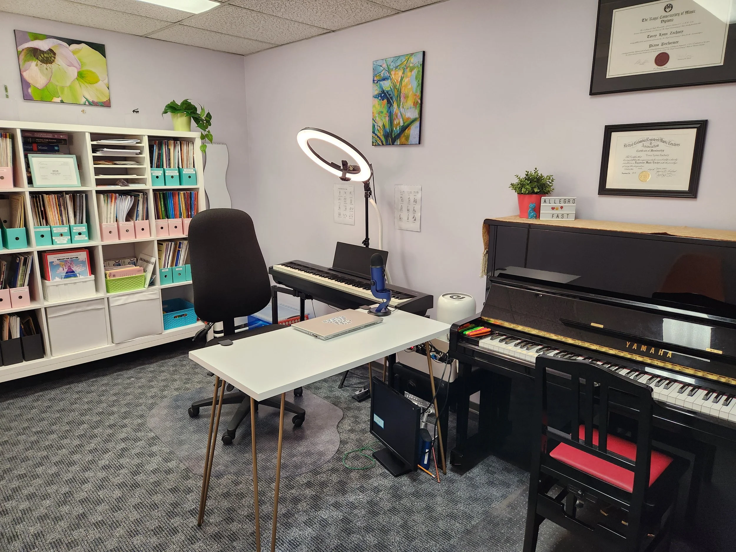 Photo of piano studio with an acoustic piano, digital piano, desk and large shelf full of sheet music