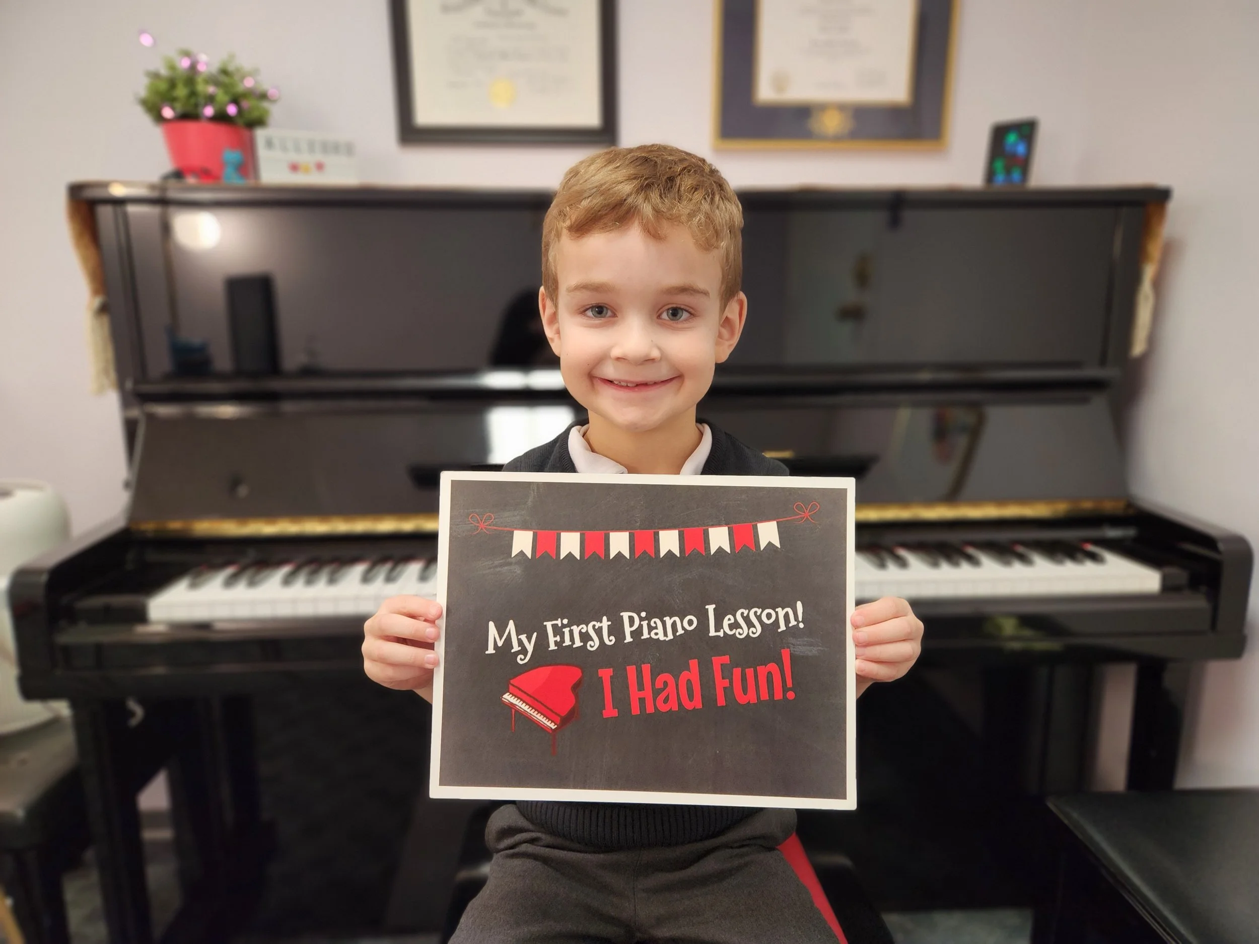 Young boy smiling, sitting in front of the piano facing the camera holding a sign that says "My First Piano Lesson! I Had Fun!"