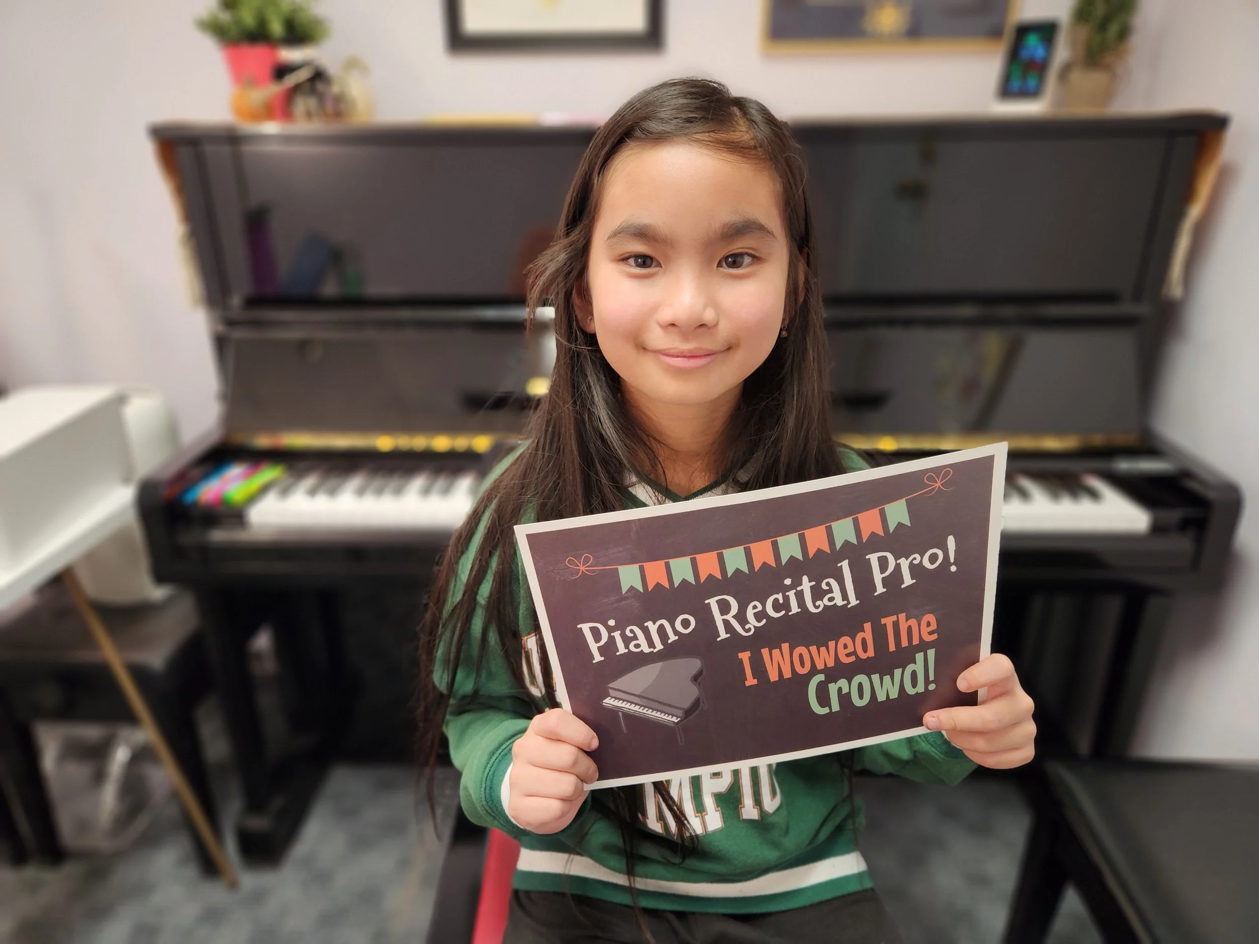 Young girl facing the camera seated in front of piano holding sign that says "Piano Recital Pro! I Wowed the Crowd!" 