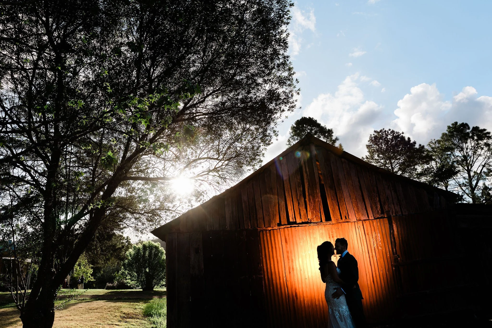 A bride and groom standing close together in front of a rustic wooden barn, backlit by the sun, with trees and a partly cloudy sky in the background. Hunter Valley NSW Wedding. David Maltby Images