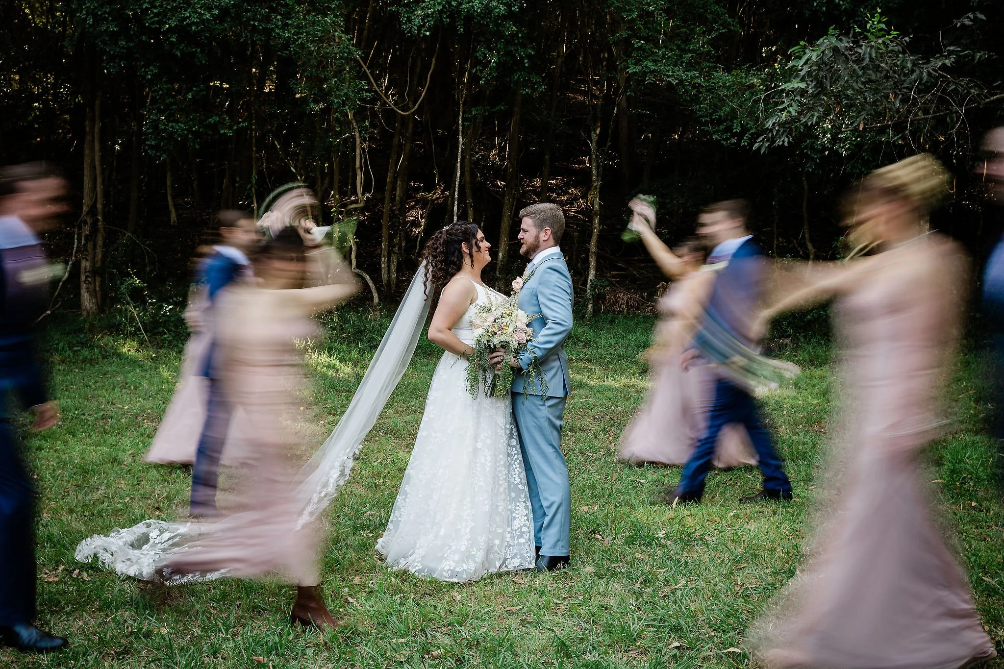 A wedding couple standing in a grassy area surrounded by trees, with blurred guests dancing around them. Hunter Valley NSW Wedding. David Maltby Images
