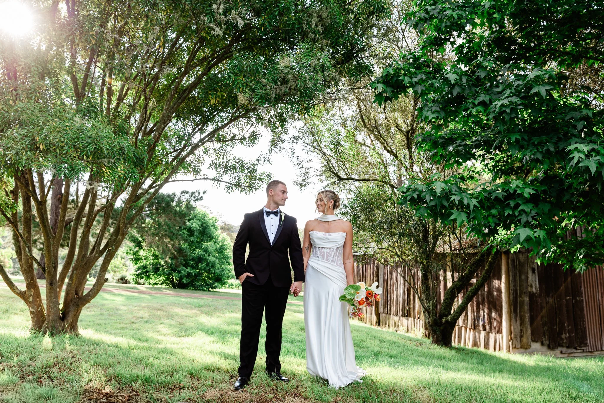 A bride and groom holding hands and looking at each other outdoors in a garden with trees and sunlight filtering through the leaves. Hunter Valley NSW Wedding. David Maltby Images