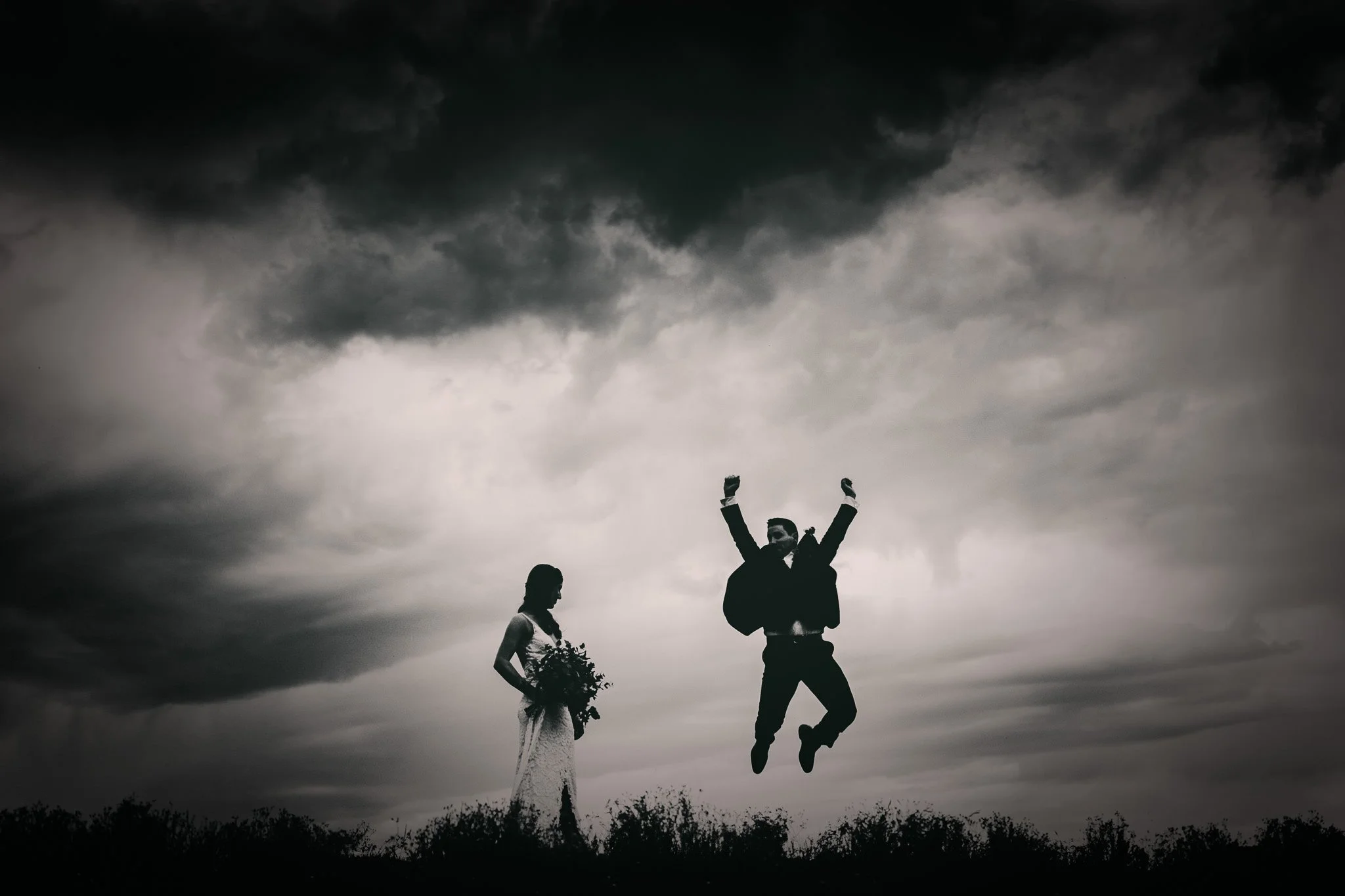 Silhouette of a bride holding a bouquet and a groom jumping in the air with arms raised under a cloudy sky. Hunter Valley NSW Wedding. David Maltby Images
