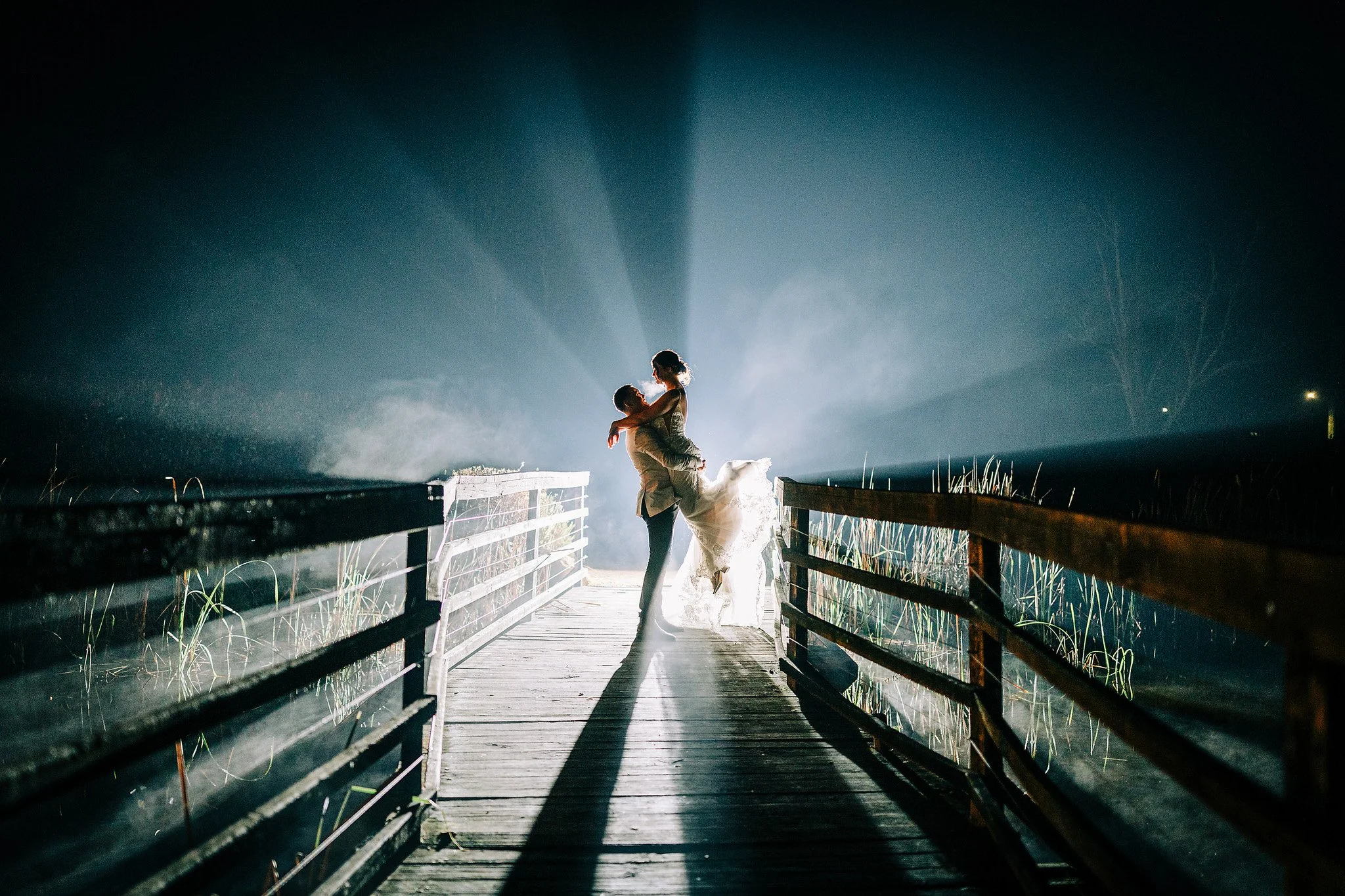 A couple dancing on a wooden dock at night with dramatic backlighting creating a silhouette effect, with water and reeds visible on either side. Hunter Valley NSW Wedding. David Maltby Images