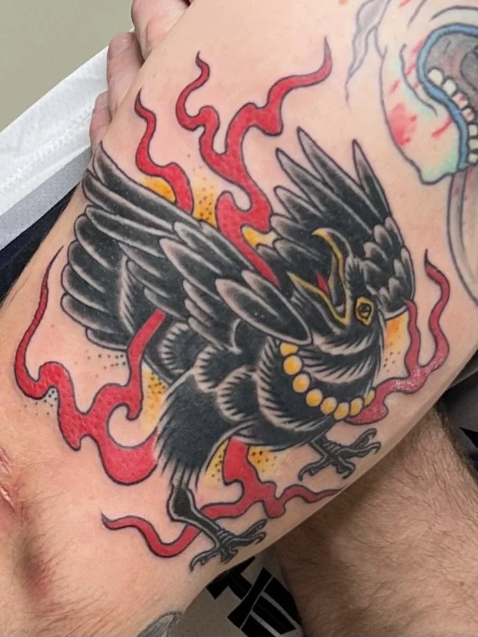 A tattoo of a black bird with spread wings, wearing a beaded necklace, with red flames surrounding it.