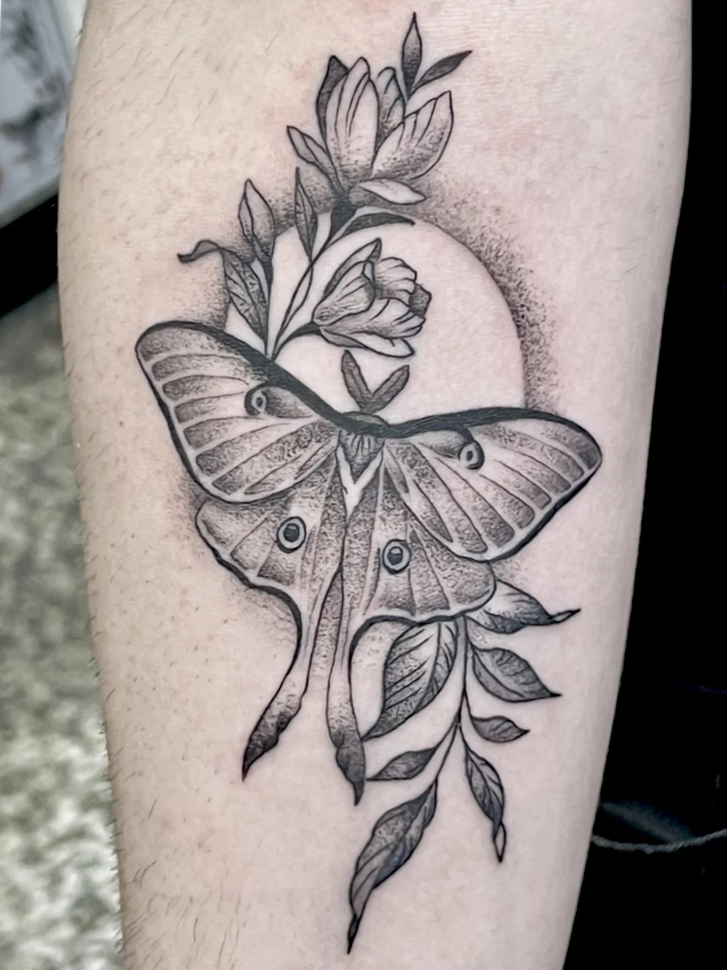 Black and gray tattoo of a butterfly with detailed wings, surrounded by leaves and flowers on a person's arm.