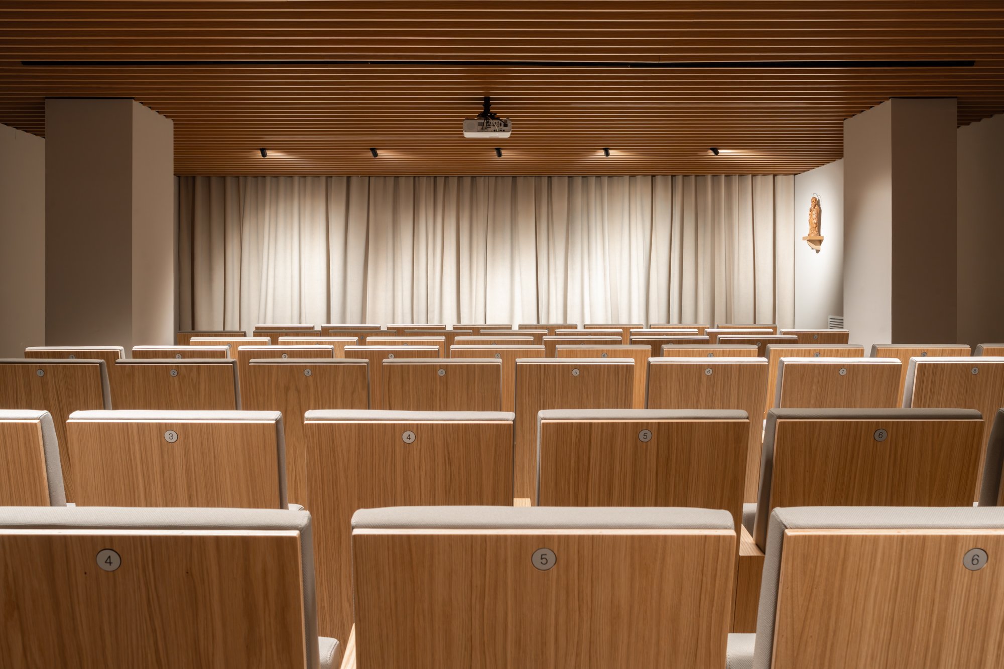 Salón de conferencias con asientos de madera y cortinas blancas en el escenario.
