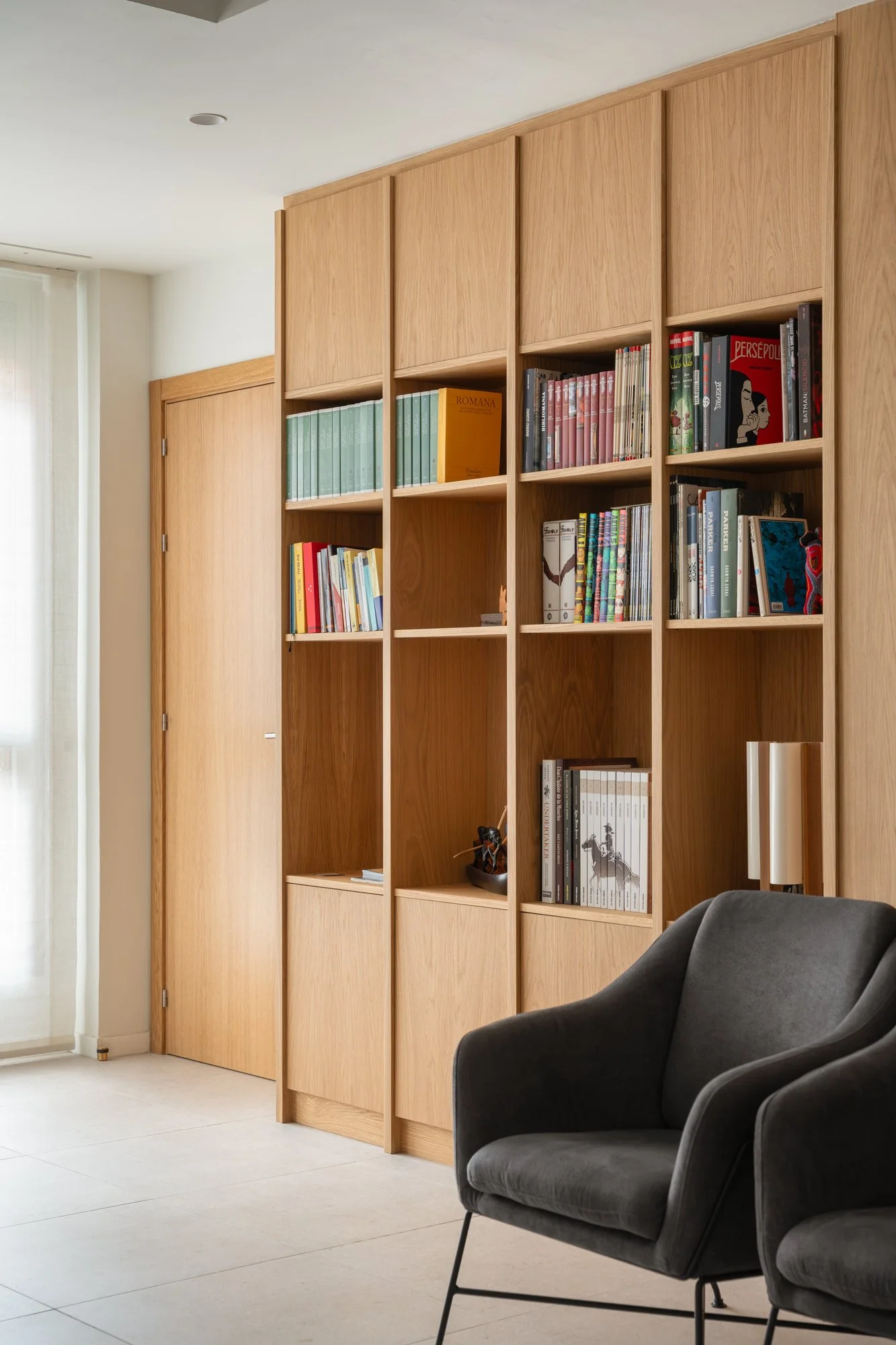 Estantería de madera con libros en una sala moderna, cerca de una puerta y una ventana con cortinas blancas, y una silla gris en primer plano.