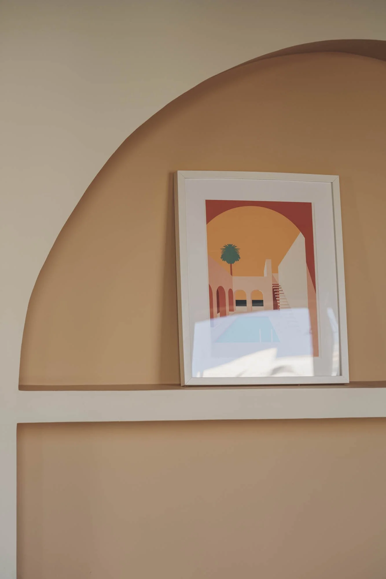 Cuadro con ilustración de un paisaje con piscina, palmera, arco y escaleras en tonos cálidos, colgado en una pared de color durazno.