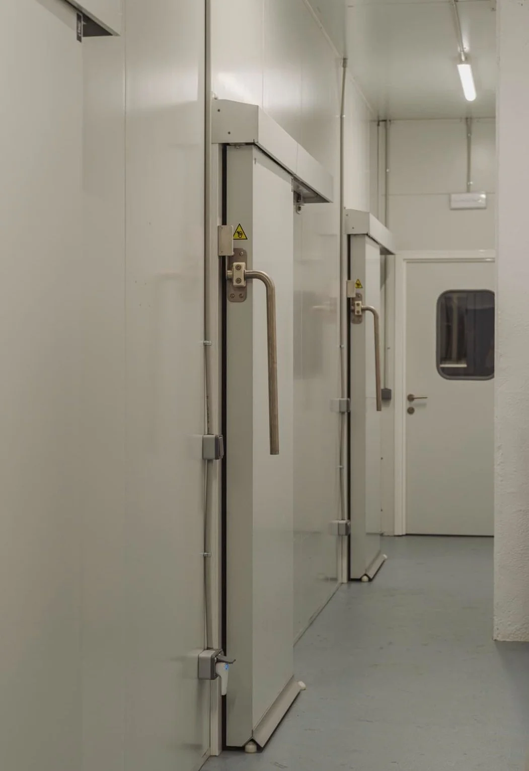 Puertas de refrigeración en un pasillo de área industrial o de laboratorio, con manijas de acero y señalización de advertencia