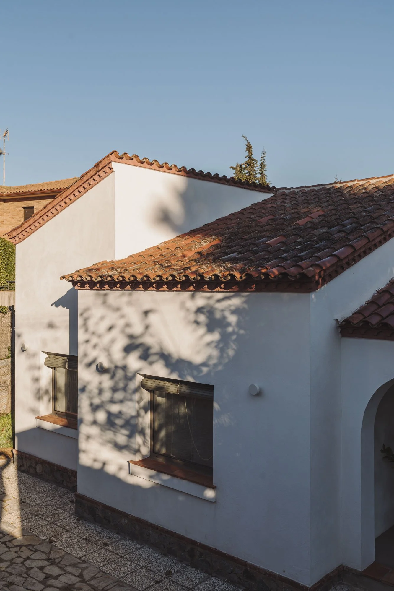 Casa con paredes blancas y techos de tejas rojas, sombra de árbol en la fachada, ventanas con persianas y entrada con arco.