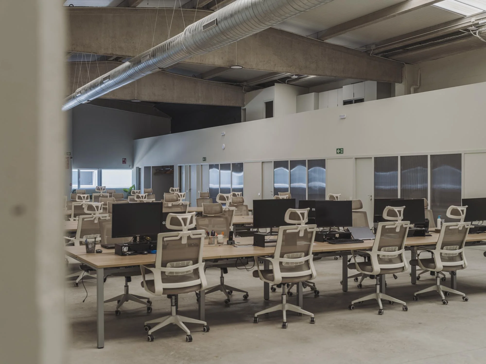Oficina vacía con escritorios y sillas, computadoras y monitores. Ventanas y paredes blancas, techo de cemento expuesto con tuberías de aire acondicionado.