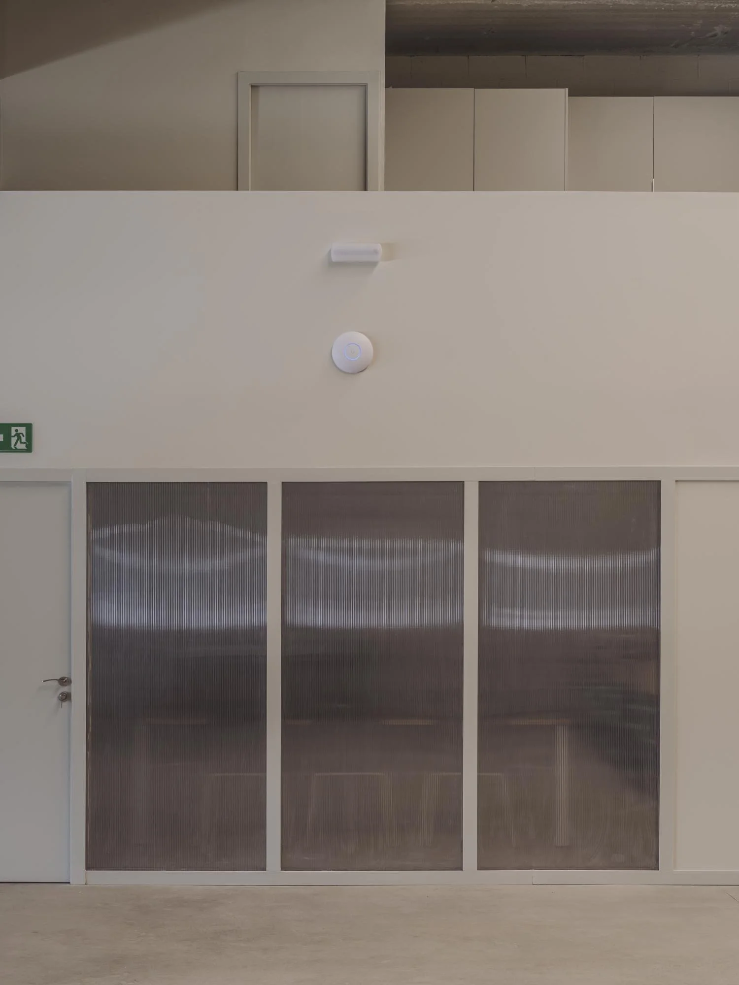 Vista de un espacio interior moderno con una pared blanca, puertas de vidrio con marco blanco y un área superior con armarios beige y una estructura en el techo.