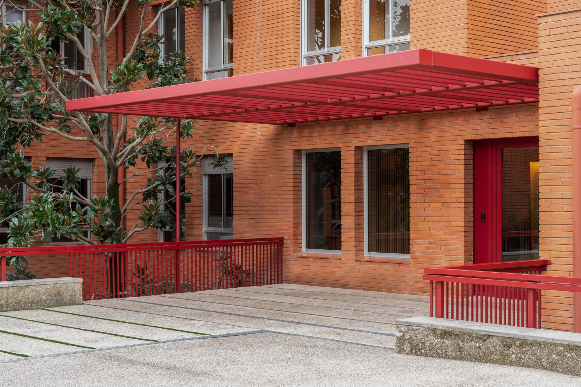Aparcamiento exterior con una estructura de techo roja y baranda del mismo color. Edificio de ladrillo con ventanas y una puerta de color rojo.