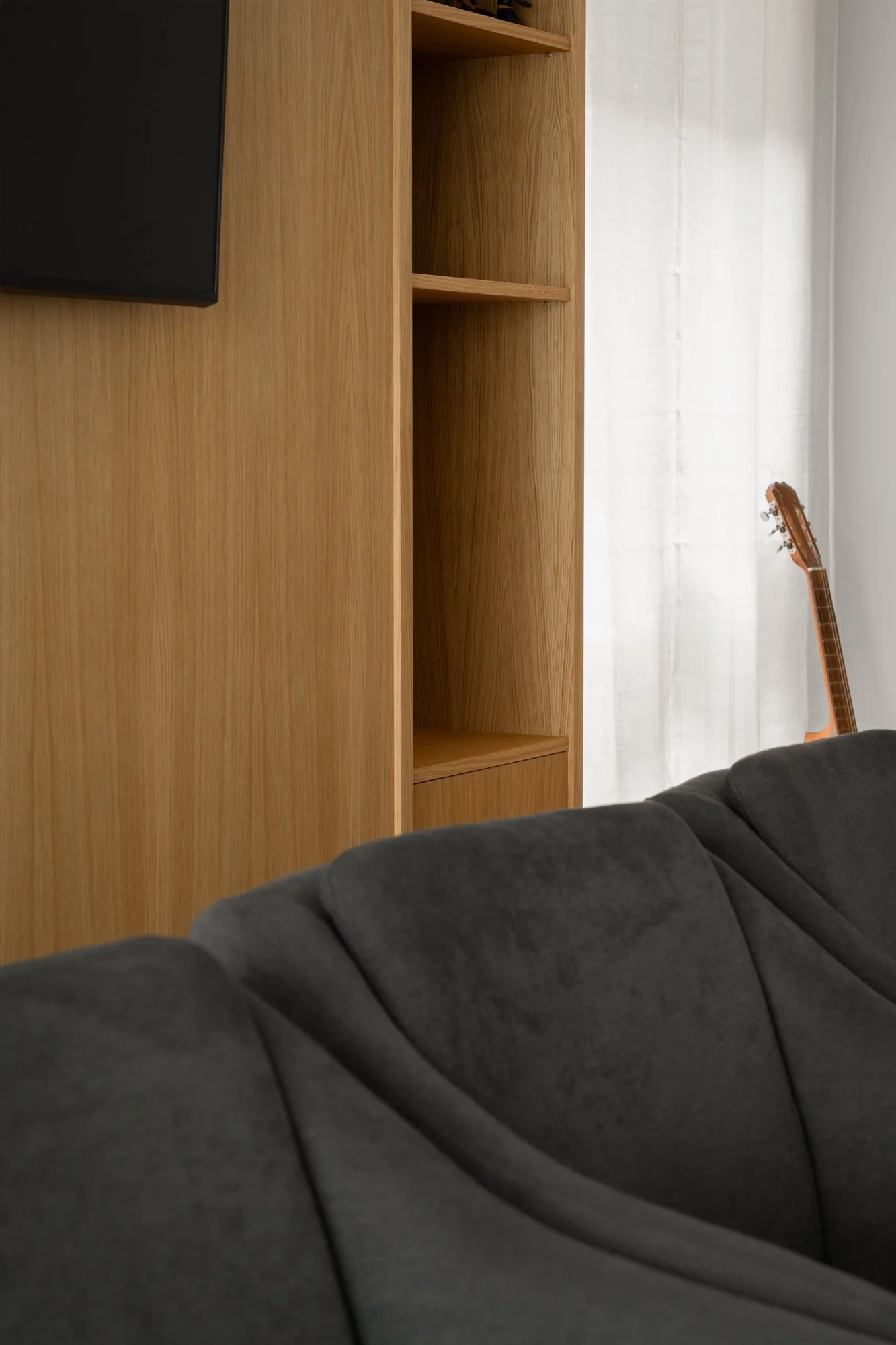 Interior de sala con pared de madera, televisor, estantería vacía y guitarra en esquina, con sofá gris oscuro en primer plano.