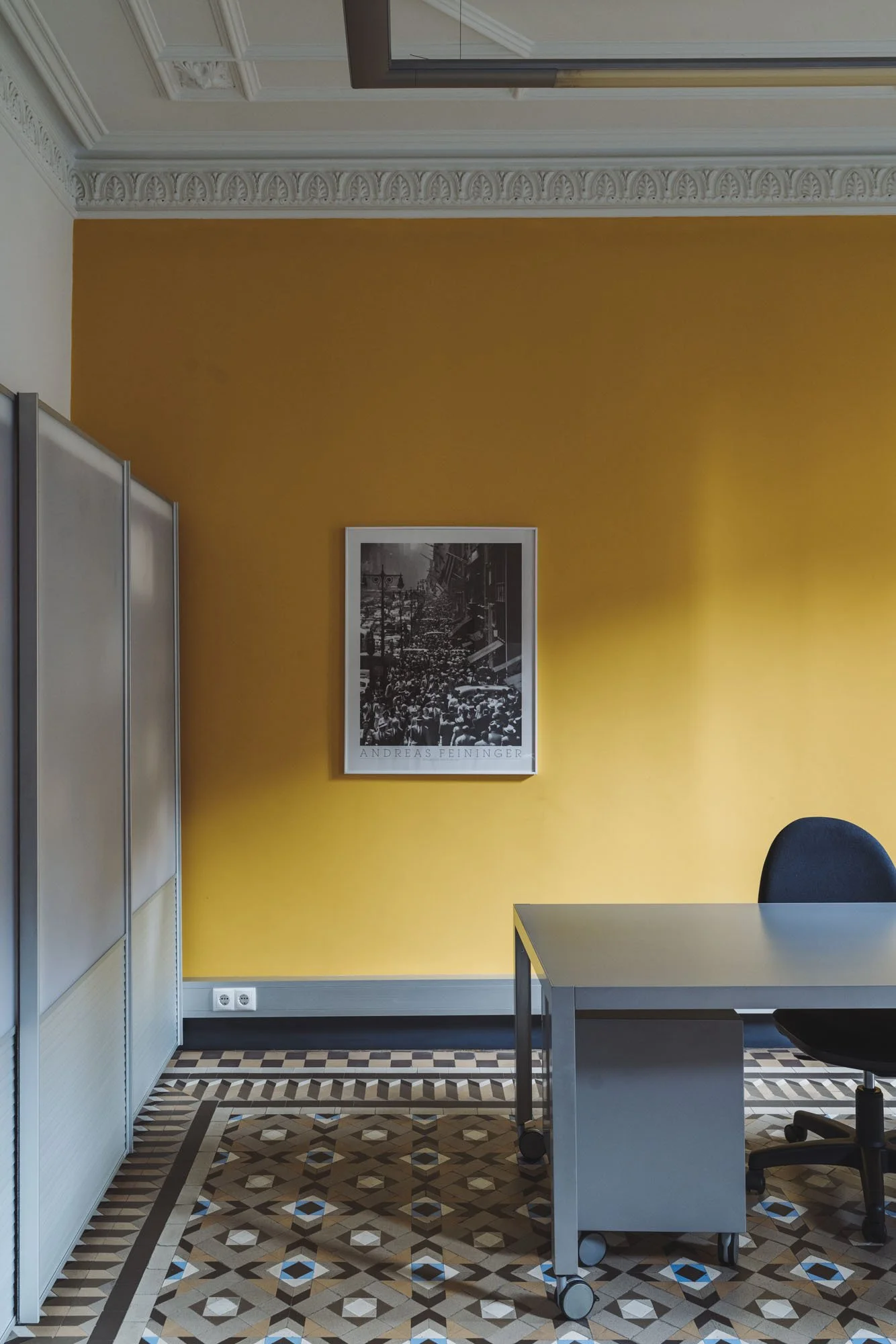 Oficina con pared amarilla, escritorio gris, silla negra, mueble metálico, cuadro en blanco y negro y piso con mosaico geométrico.