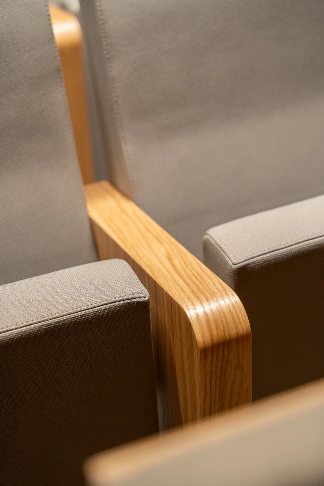 Asientos con respaldo y brazos de madera clara en una sala con telón de fondo gris.