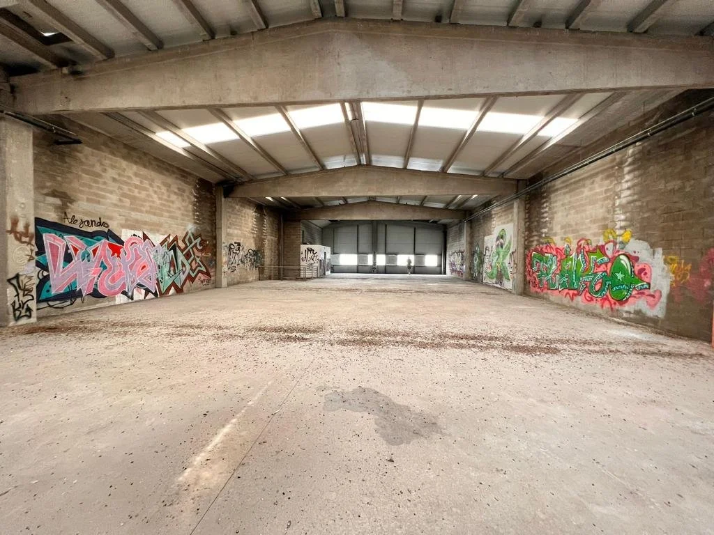 Interior de un espacio amplio con paredes de ladrillo y graffiti en ellas, suelo de cemento y techo de estructura metálica.