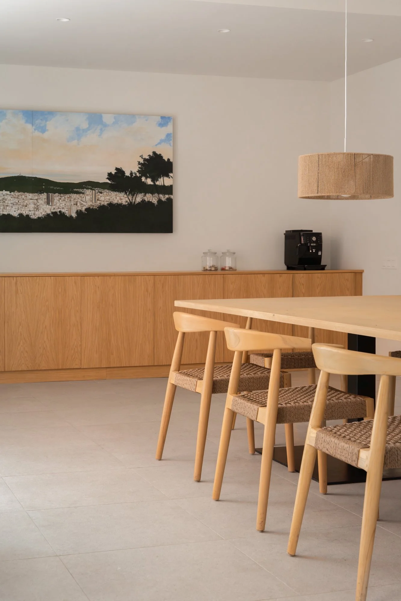Sala de comedor moderna con mesa de madera y sillas de madera con asientos de fibra, un cuadro en la pared con paisaje y un dispensador de café en la esquina