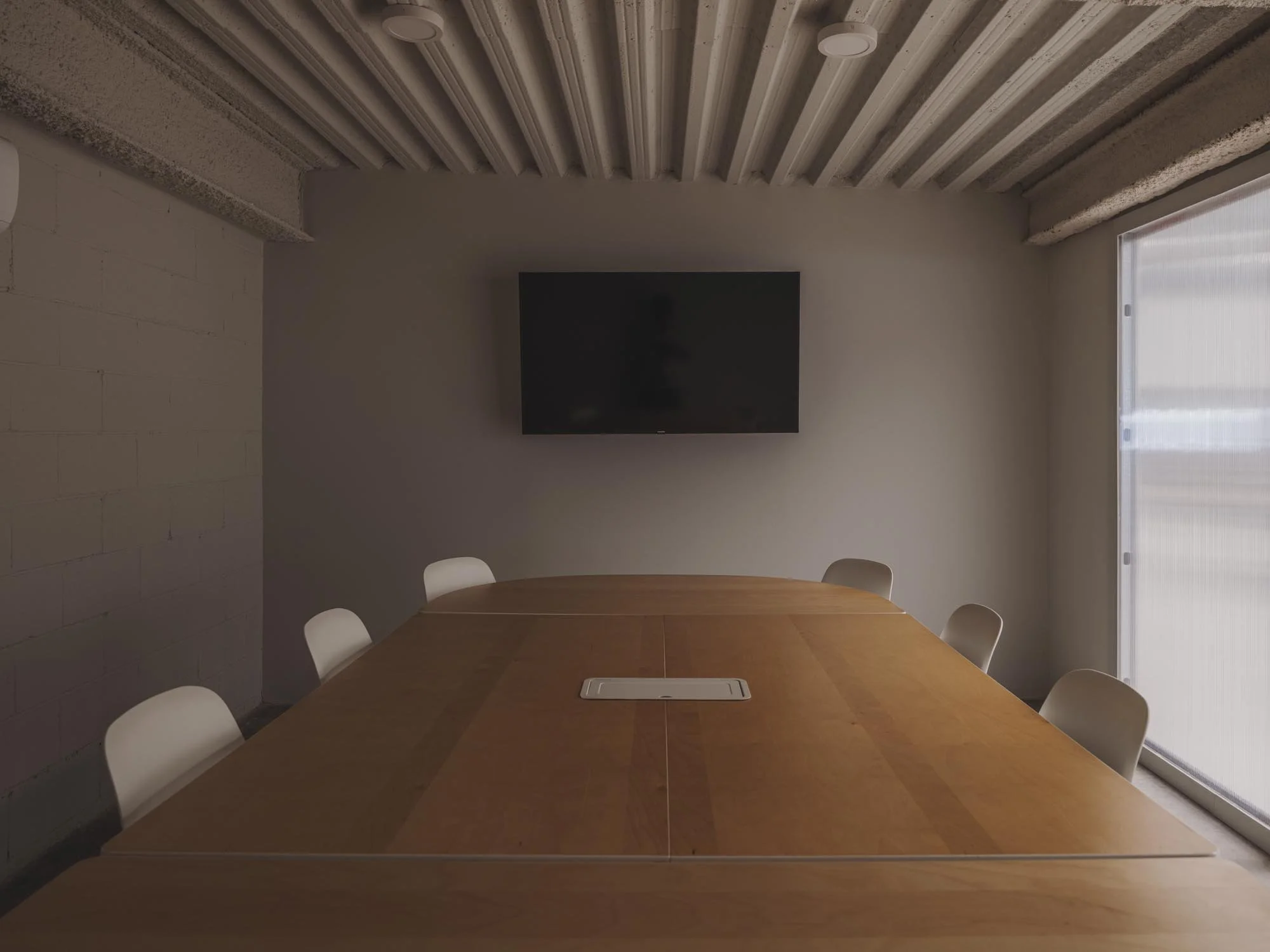 Sala de conferencias con mesa de madera, seis sillas blancas, televisión en la pared y ventanas con cortinas blancas.