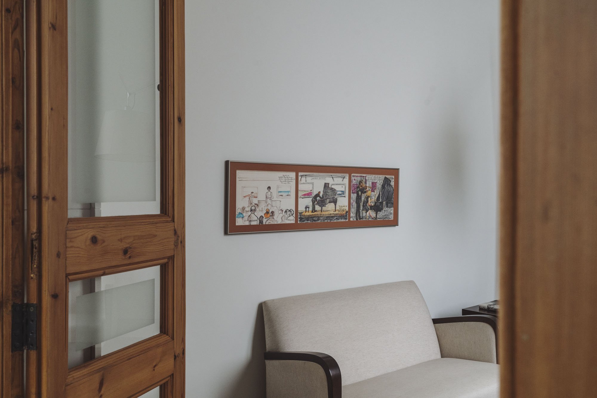 Vista de una sala con un sillón beige, un cuadro en la pared y una mesa de noche con algunos objetos, vista desde una puerta de madera entreabierta.