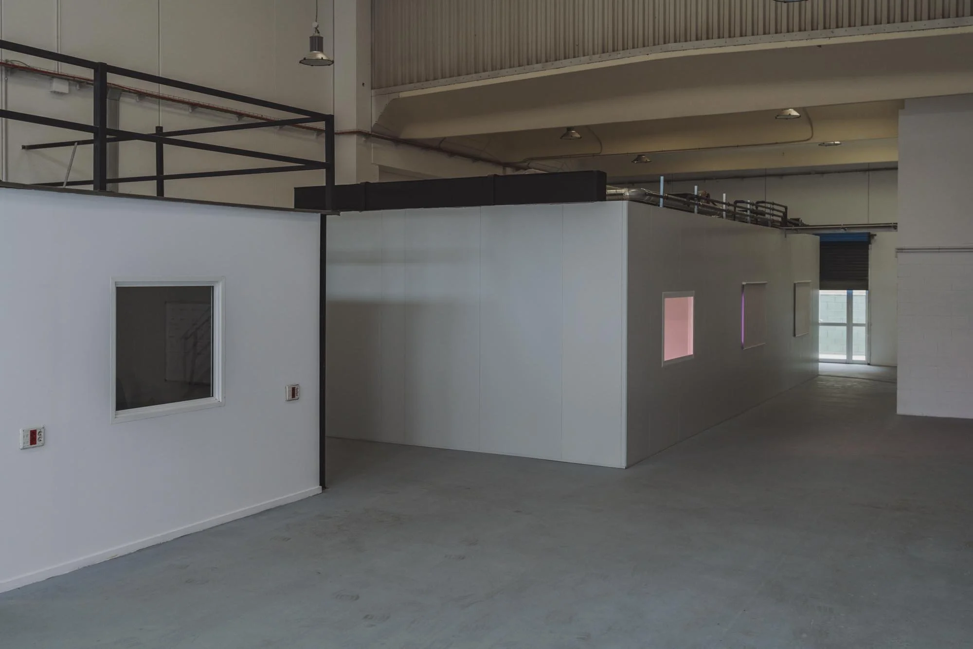 Interior de un espacio industrial o de oficina con paredes blancas, ventanas pequeñas con cristales y una puerta de vidrio en el fondo, piso de concreto y techo alto con luces instaladas.