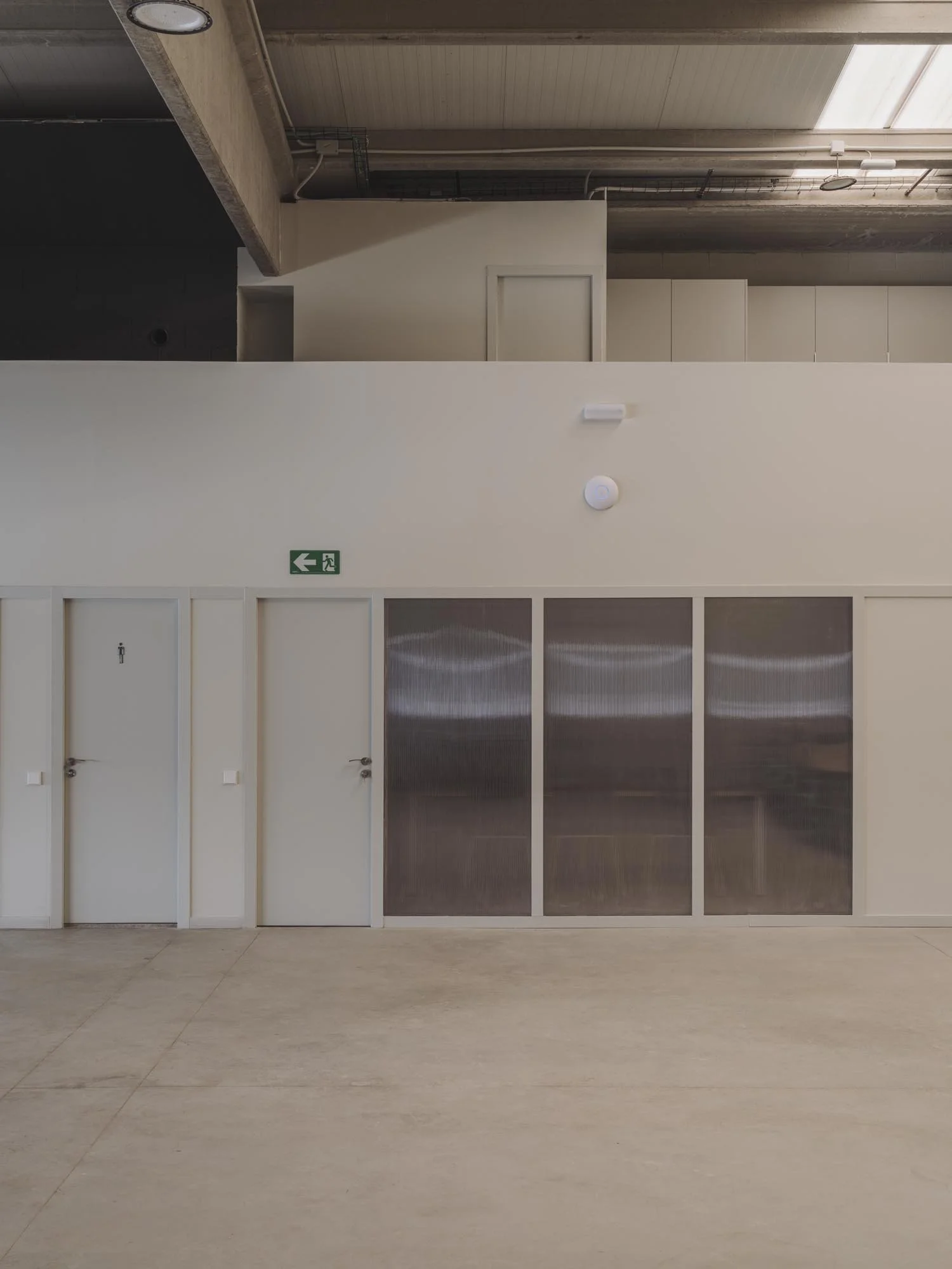 Vista de un muro con un letrero de salida de emergencia, un puerta de baño para hombres, una puerta con un cartel en blanco, y ventanas de vidrio, en un espacio interior con suelo de concreto y techo de madera.