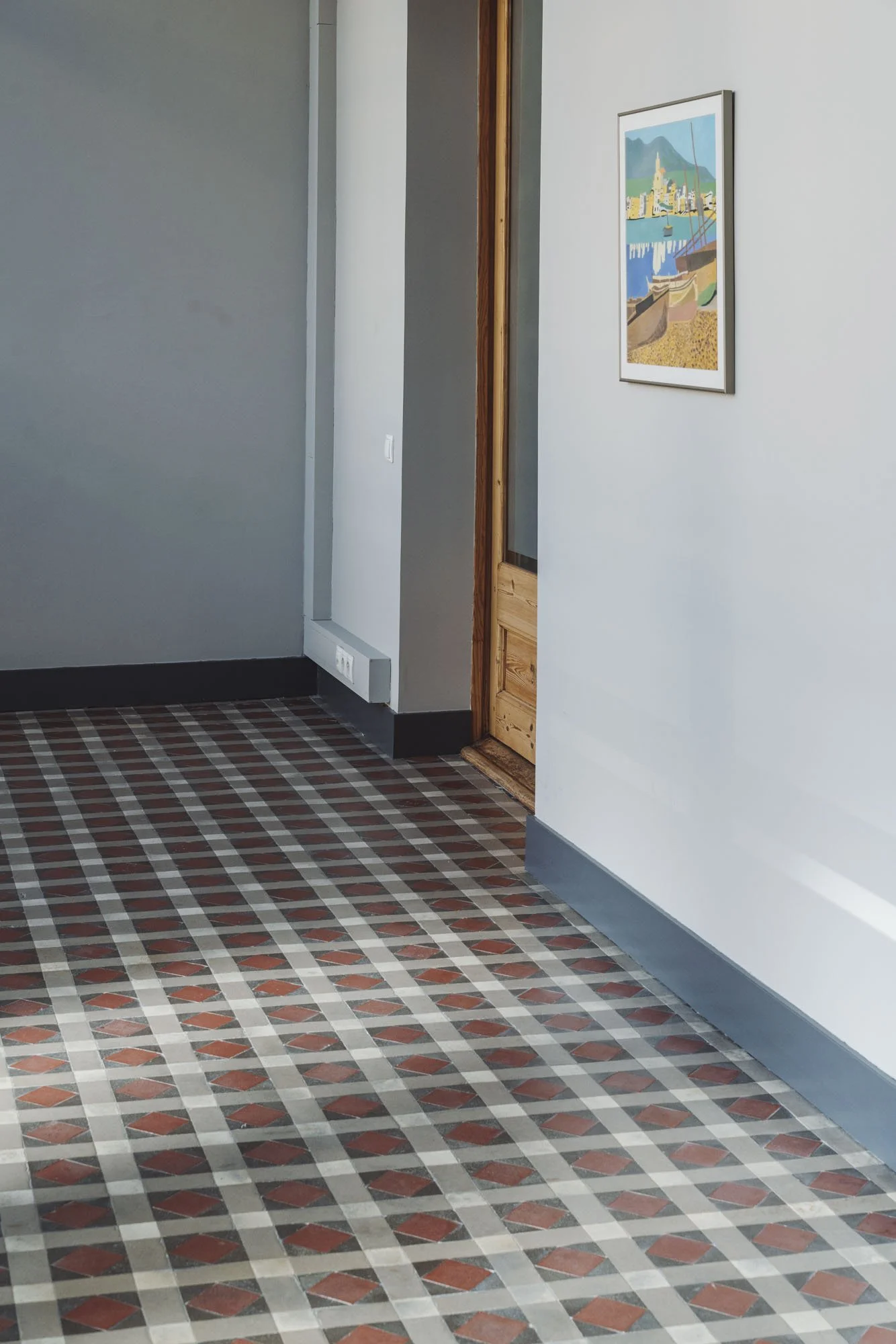 Entrada con piso de baldosas con patrón geométrico en tonos marrones y beige, pared blanca a la derecha con cuadro enmarcado y vista de puerta de madera a la izquierda.