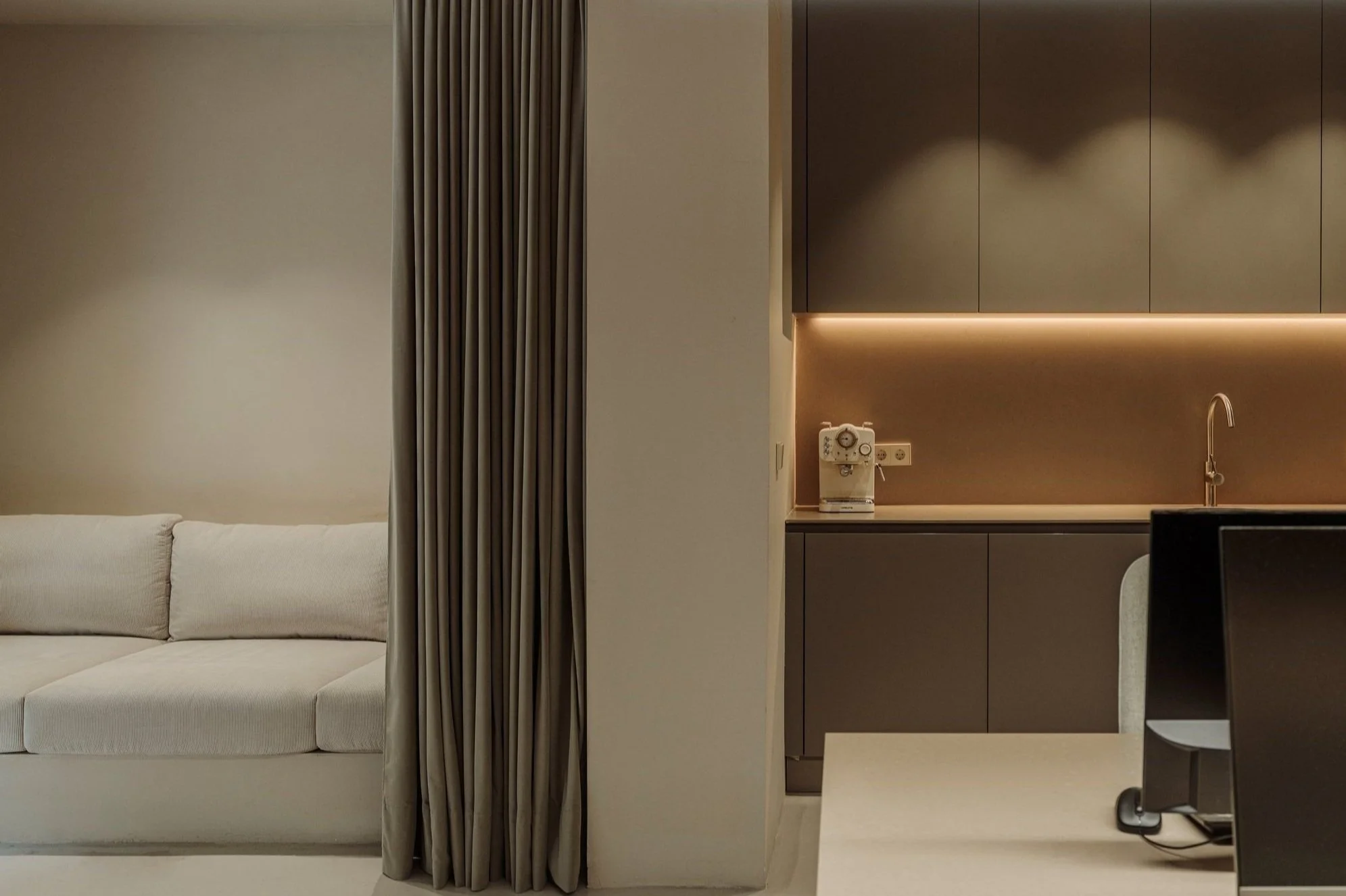 Sala de estar moderna con sofá blanco y cortinas beige a la izquierda, área de cocina con gabinetes grises, encimera beige, cafetera y lavabo, en un diseño minimalista y elegante