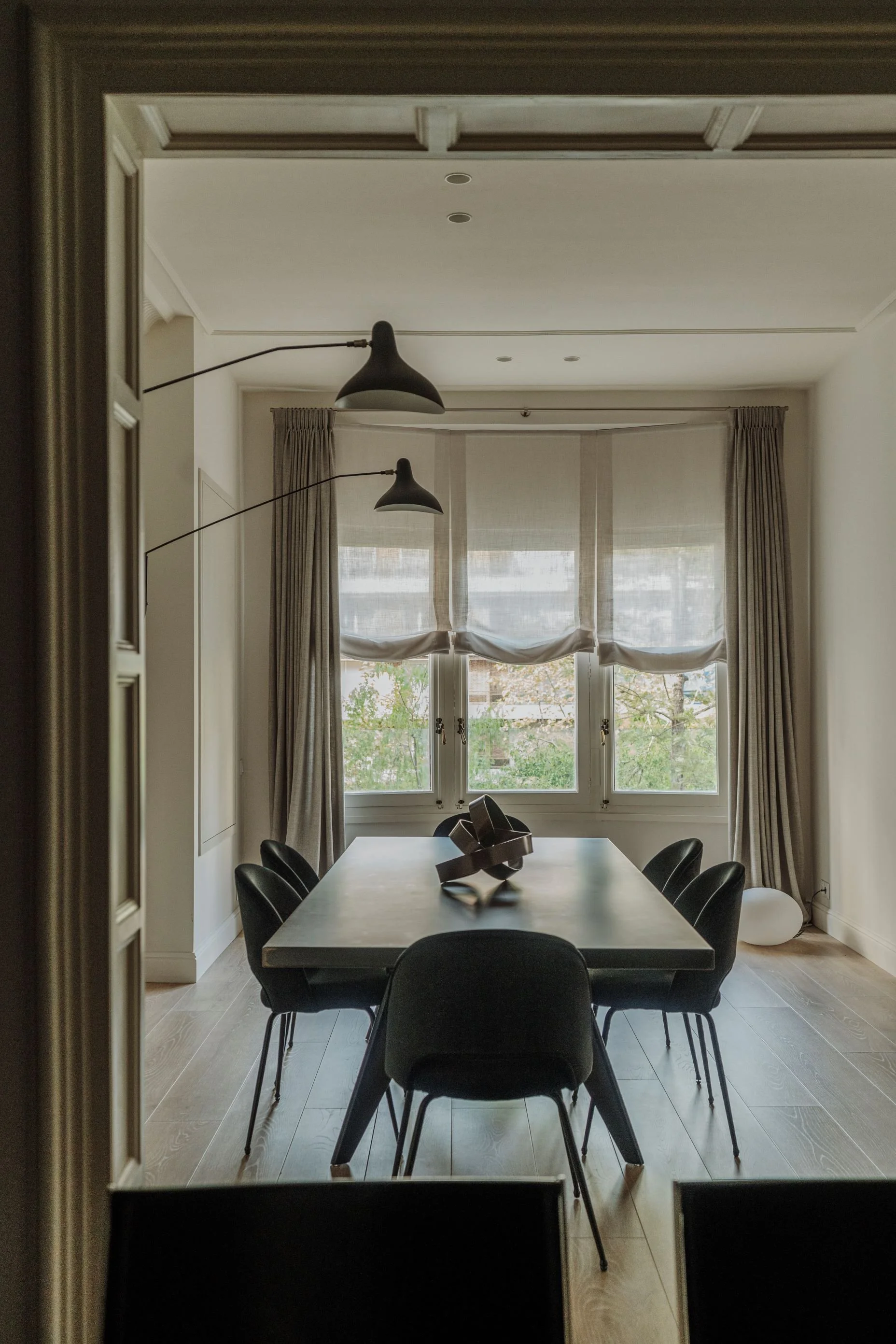 Sala de comedor moderna con mesa grande de madera y ocho sillas negras, cerca de ventanas grandes con cortinas beige y vistas de árboles, decorada con lámparas de techo negras y una escultura en el centro de la mesa.