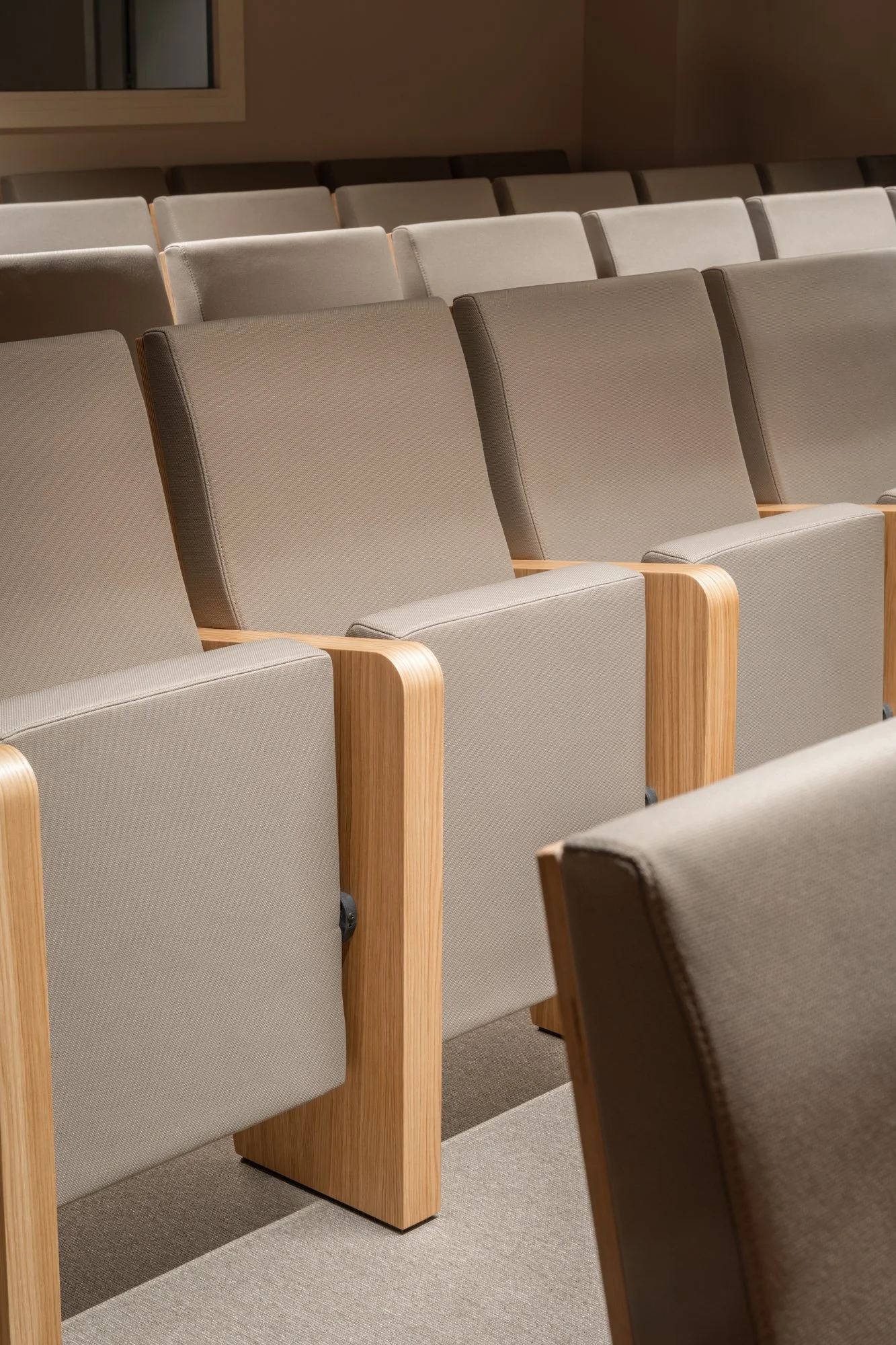 Filas de asientos en una sala vacía, con estructura de madera y respaldos y asientos tapizados en tela beige.