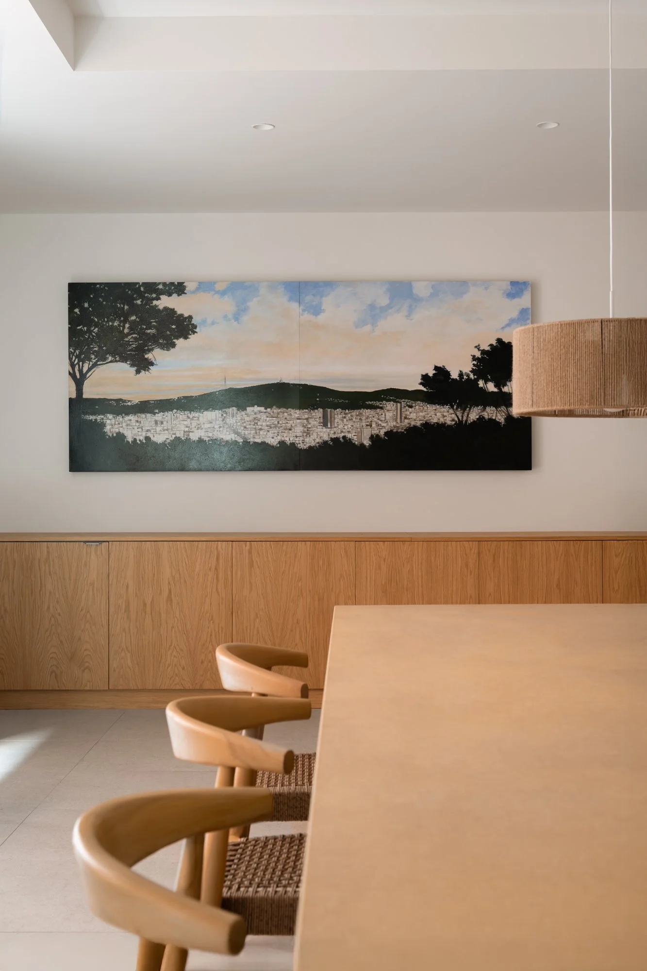 Una sala moderna con un cuadro grande de un paisaje urbano y árboles, mesas de madera y sillas de madera en primer plano.