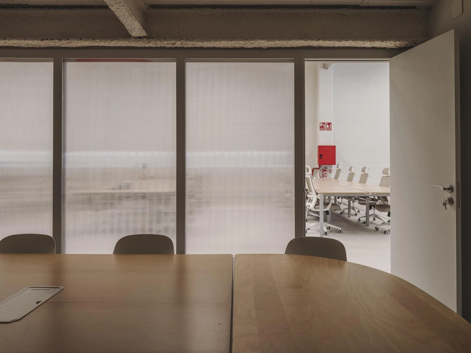 Sala de reuniones vista a través de un acrílico con vistas a otra sala con sillas y mesas blancas, y una pared con extintor rojo.