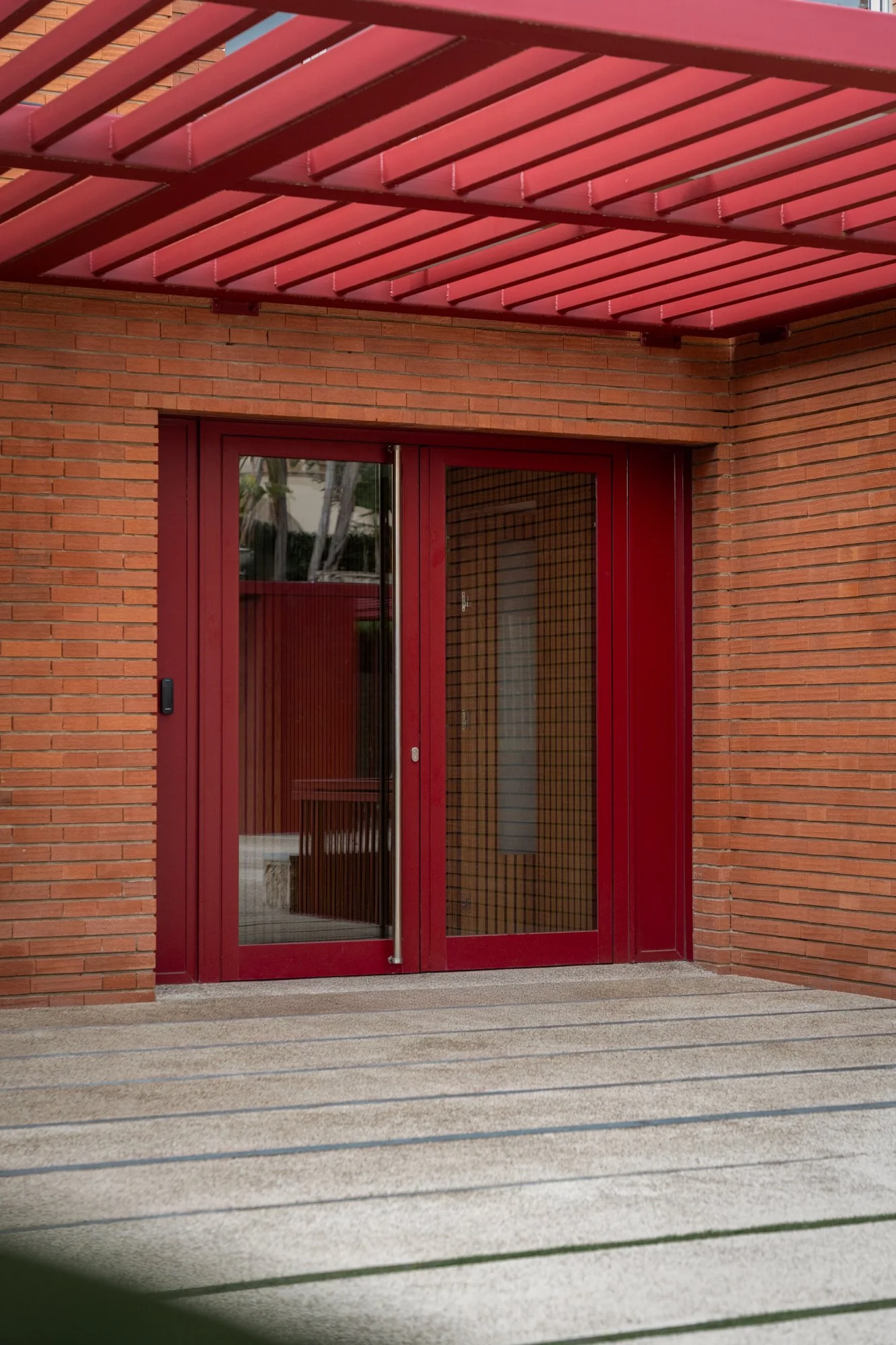 Puerta de entrada de cristal con marco rojo, rodeada de paredes de ladrillo, con una estructura de protección de metal roja en el techo y un suelo de baldosas de colores claros.