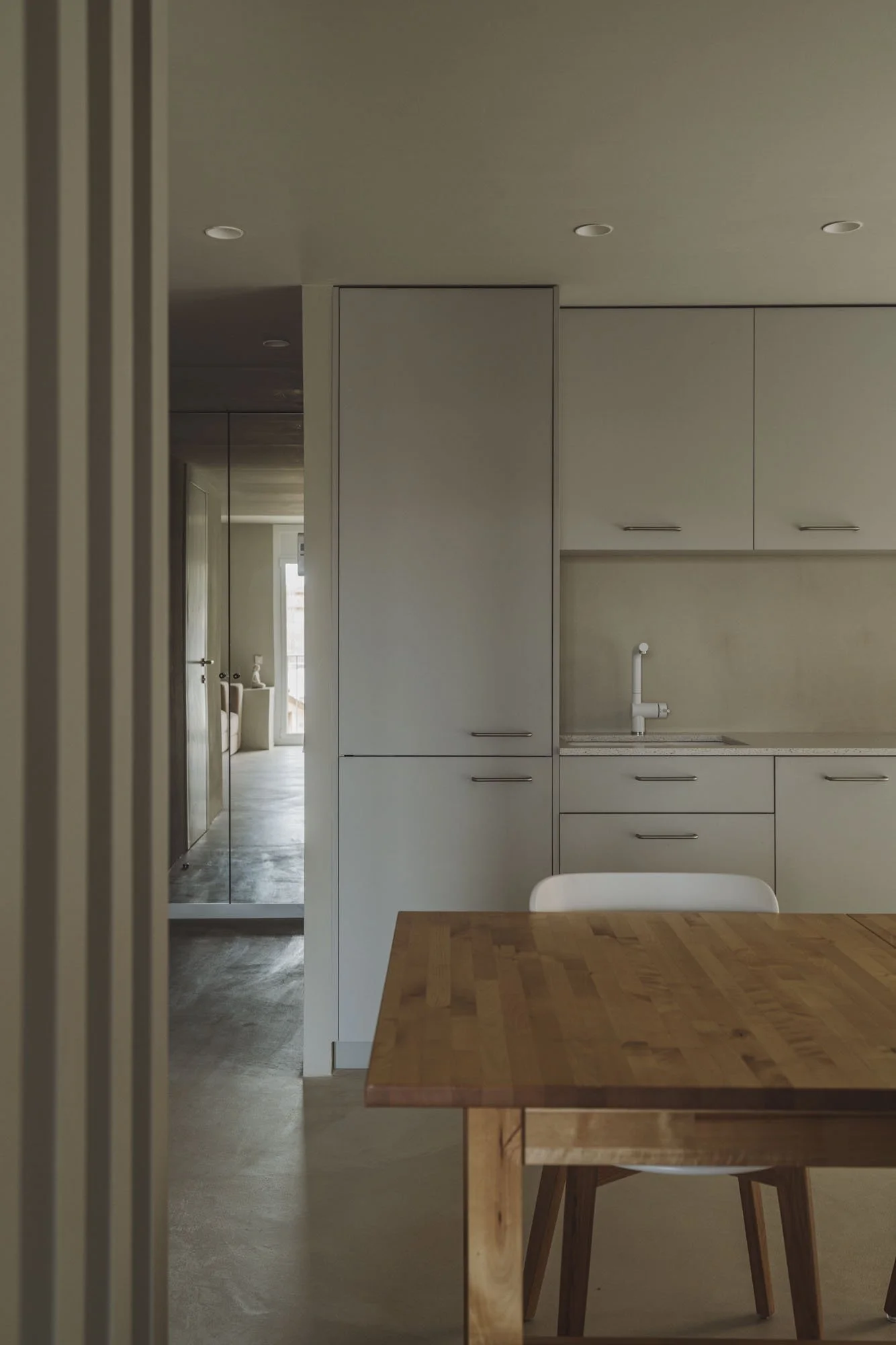 Vista parcial de una cocina moderna con gabinetes blancos, fregadero y una mesa de madera en primer plano. Se observa un pasillo con espejo y una sala al fondo con ventanas grandes.