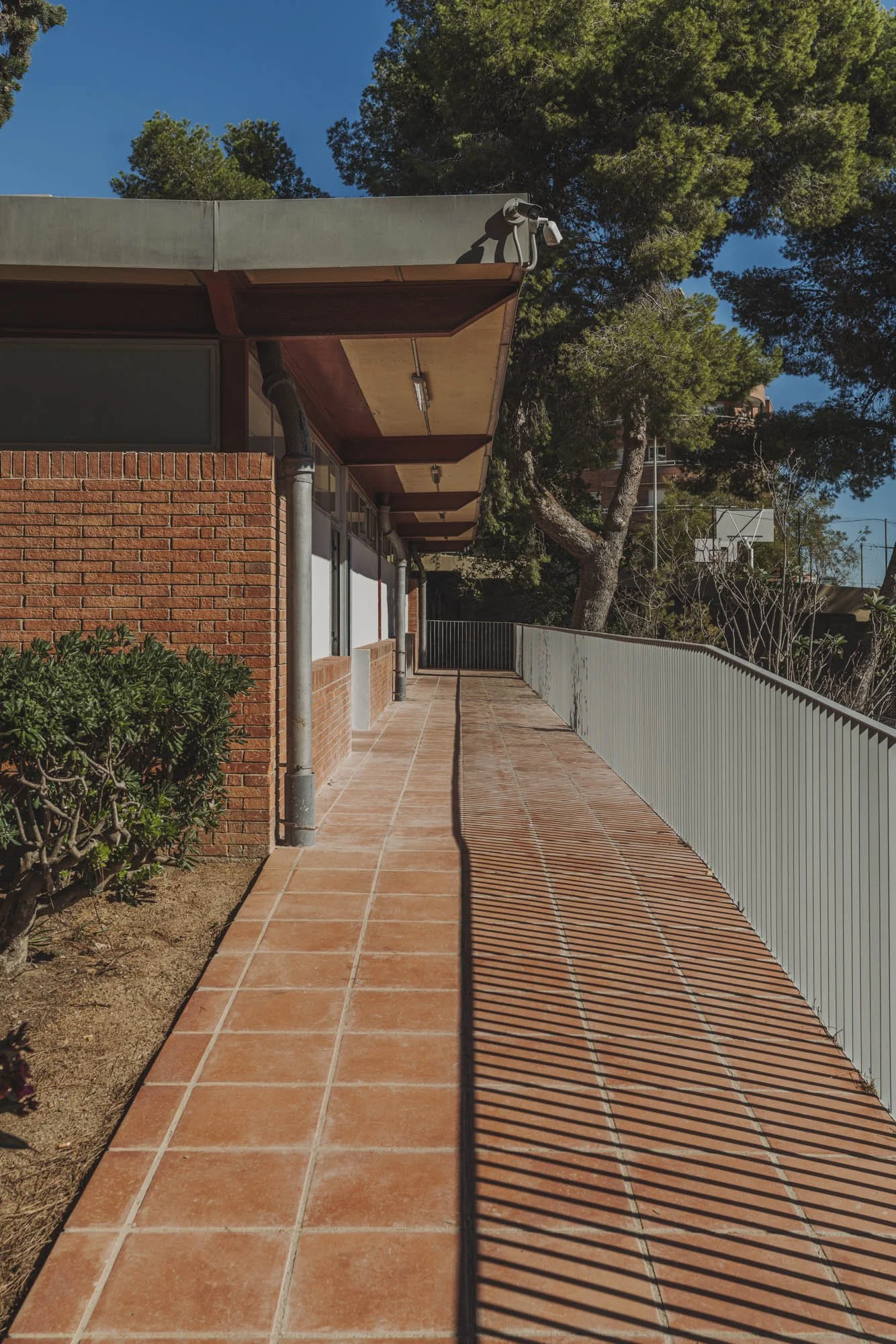 Pasillo exterior de una edificación con balcón, en un día soleado, con árboles verdes y un cielo azul en el fondo.