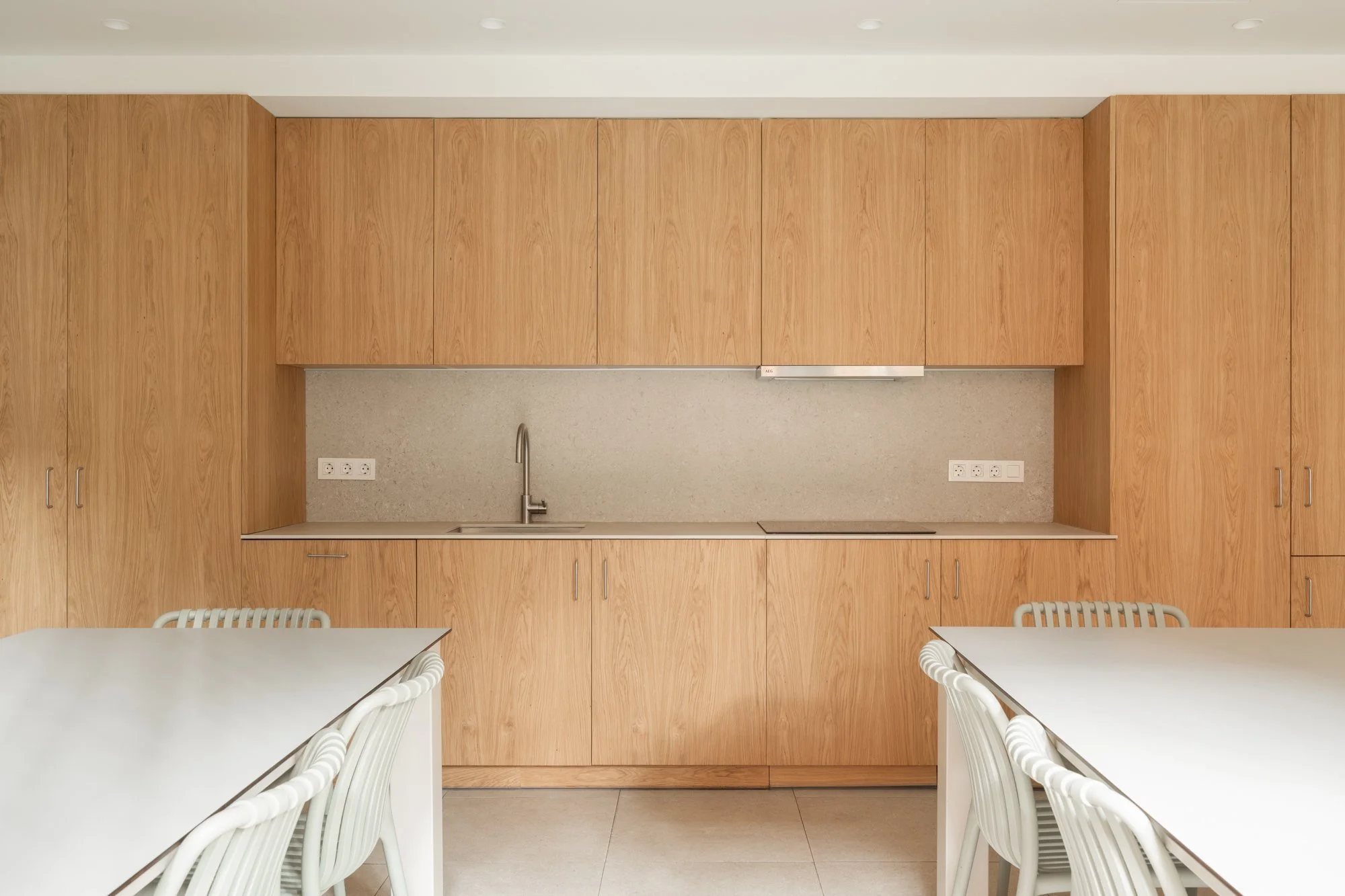 Cocina moderna con gabinetes de madera, fregadero y mesas blancas con sillas.