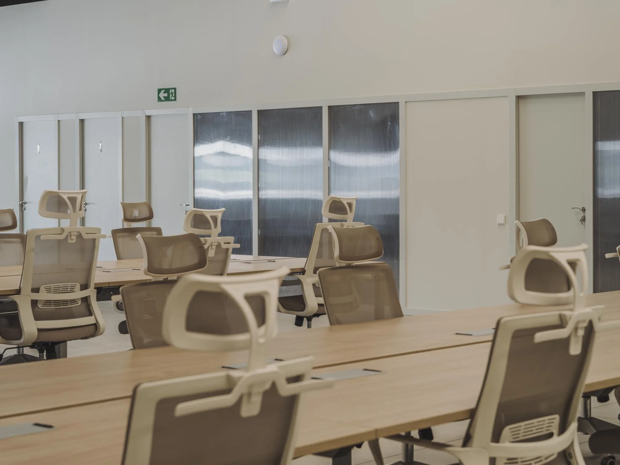 Sala de reuniones moderna con mesas de madera y sillas ergonómicas, paredes blancas y puertas con paneles de vidrio; señal de salida de emergencia en la pared.
