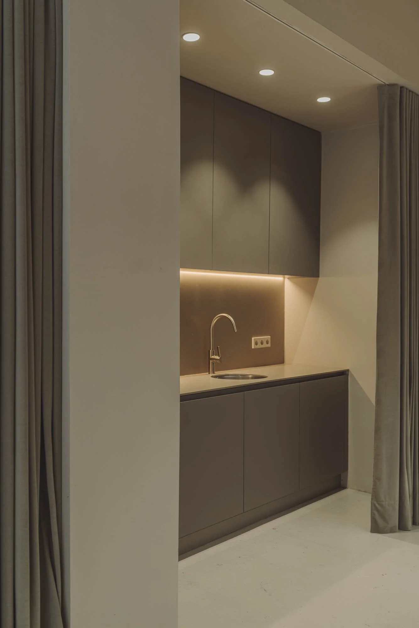 Cocina moderna con gabinetes grises y fregadero, iluminación empotrada y cortinas beige a la derecha.