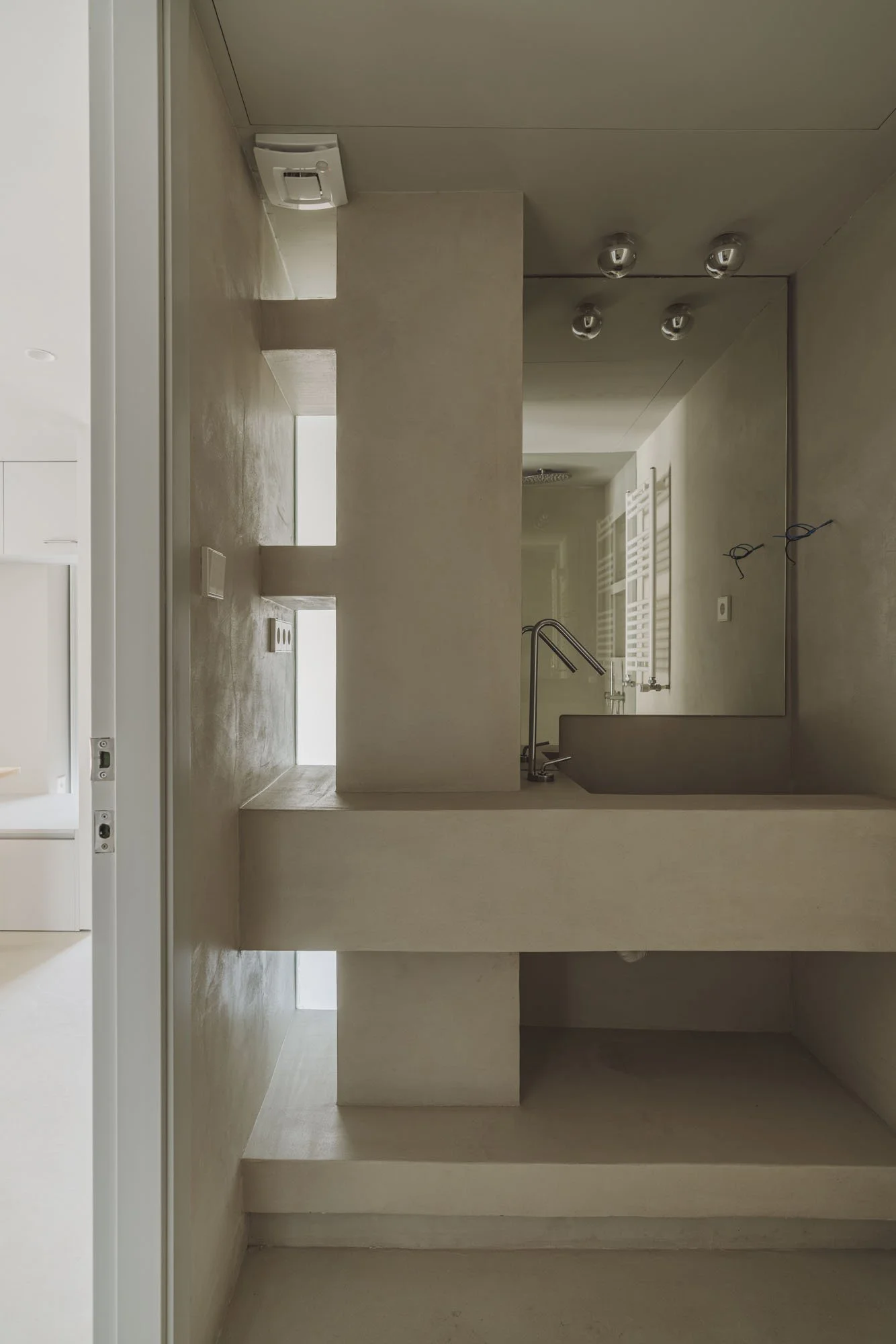 Interiores minimalistas con paredes de color beige y una ventana estrecha que deja entrar luz natural, incluyendo un fregadero de cocina con grifo y luces en el techo.