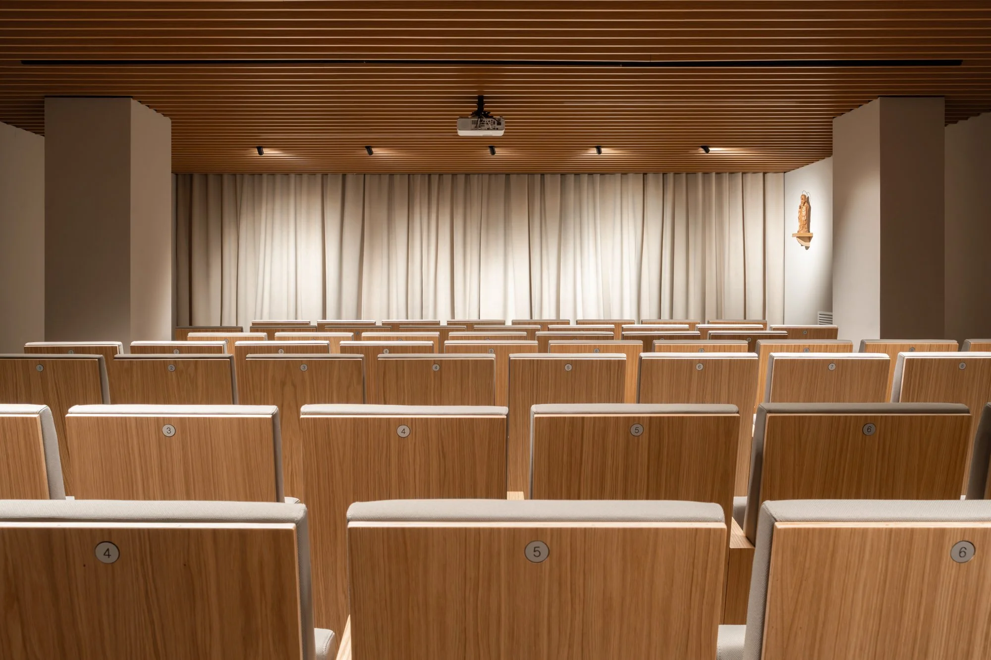 Salón de conferencias con filas de sillas de madera y cojín gris, cortinas blancas en el fondo para un escenario, techo de madera con iluminación empotrada y proyector en el centro.