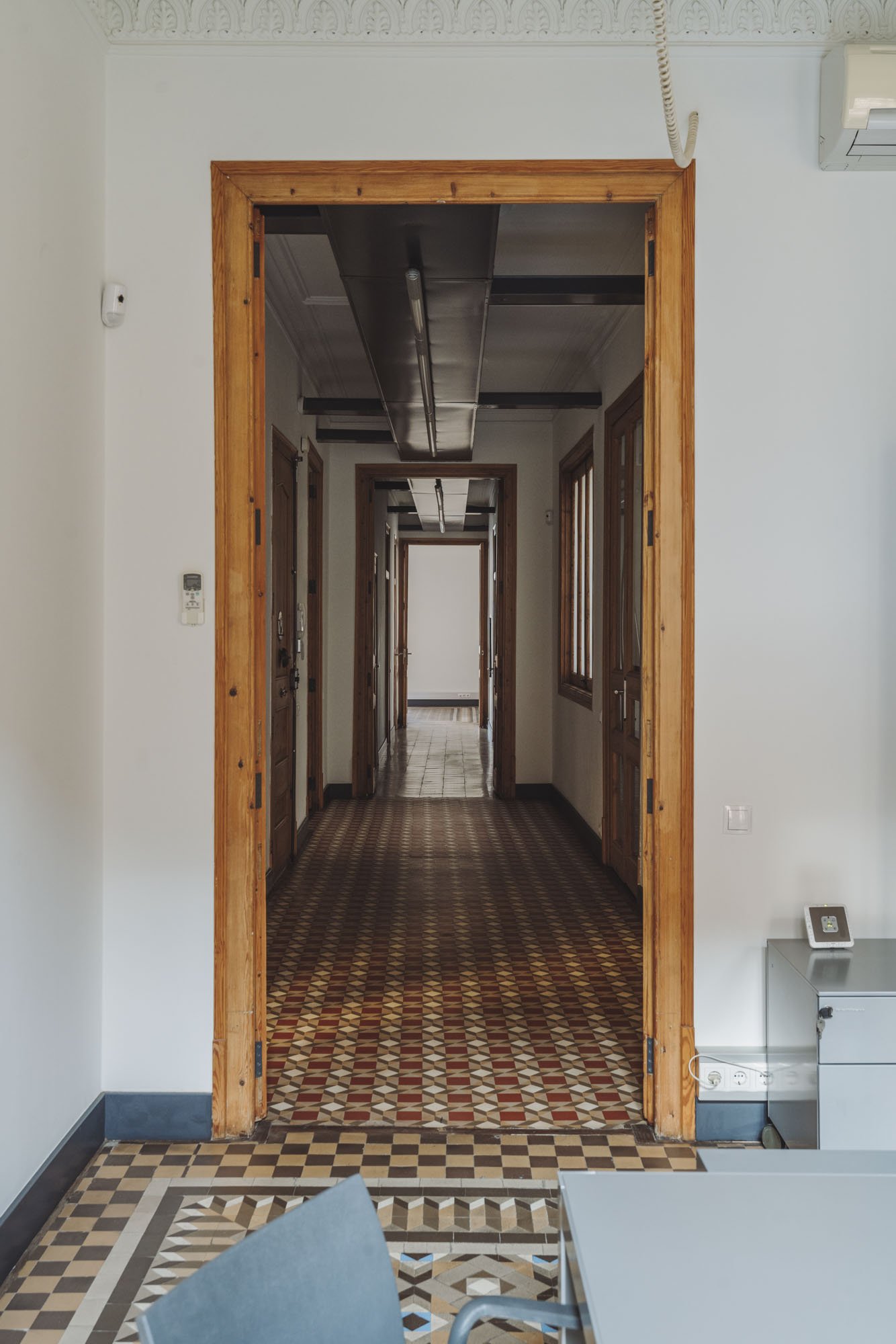 Vista de un pasillo largo visto desde una habitación con suelo de baldosas cuadradas decoradas. La puerta de la habitación tiene marco de madera. El pasillo también tiene paredes con puertas de madera y ventanas con rejas, y techo con canaletas metál